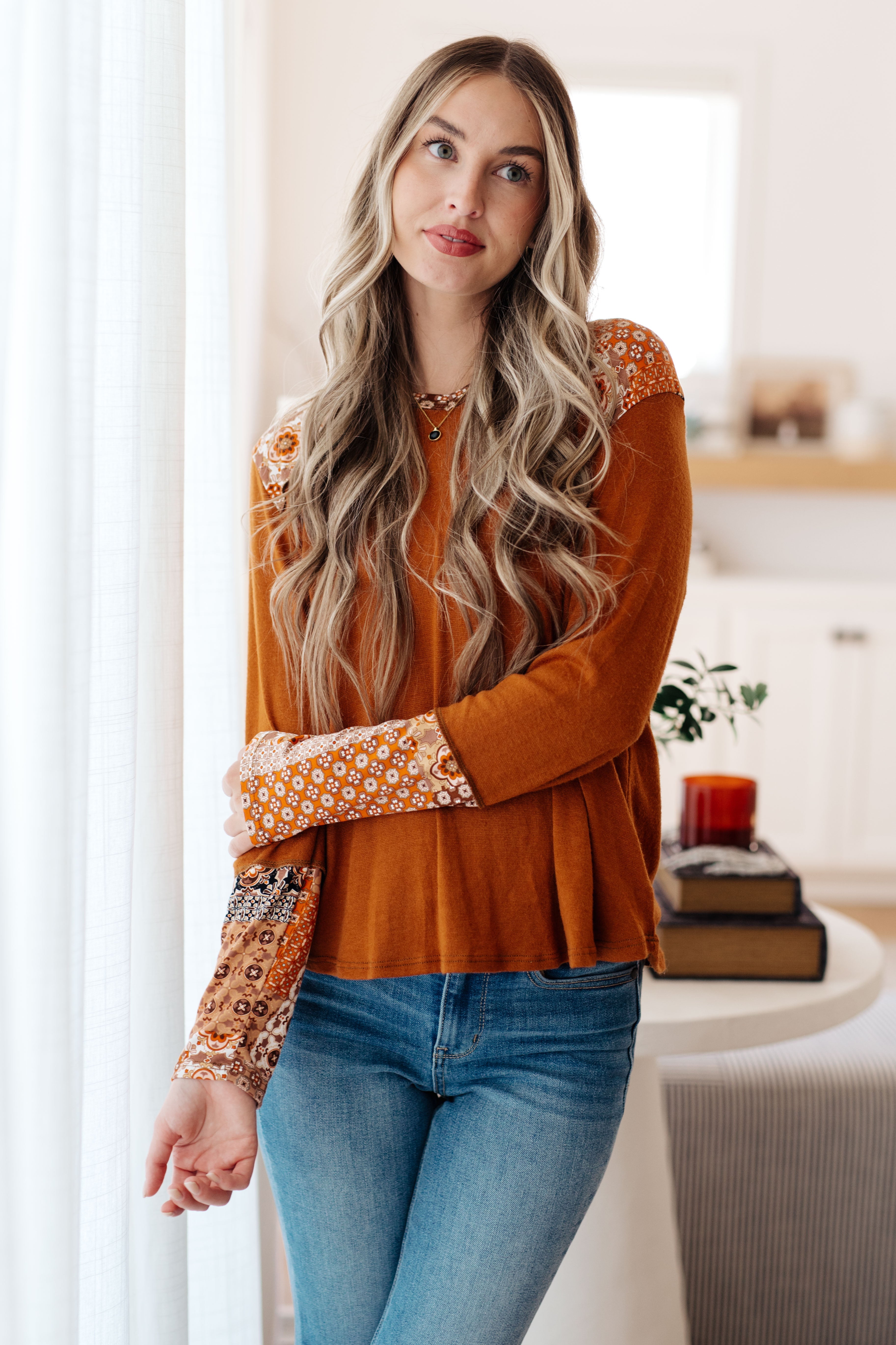 Woman wearing rust-colored mixed print hoodie with floral patchwork sleeves paired with light wash jeans indoors