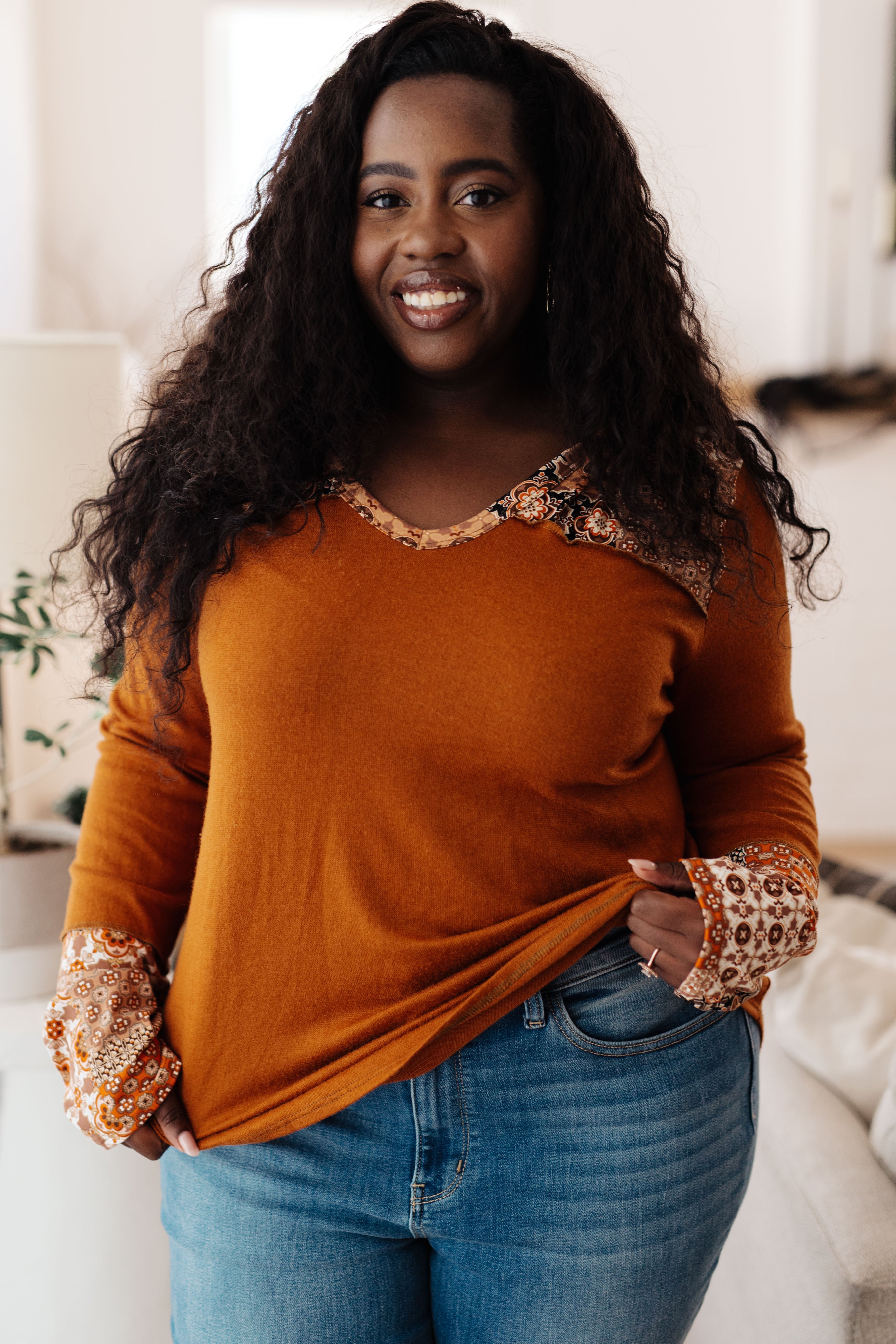 Smiling plus-size woman wearing rust mixed print hoodie with patterned trim and light blue jeans in cozy indoor setting