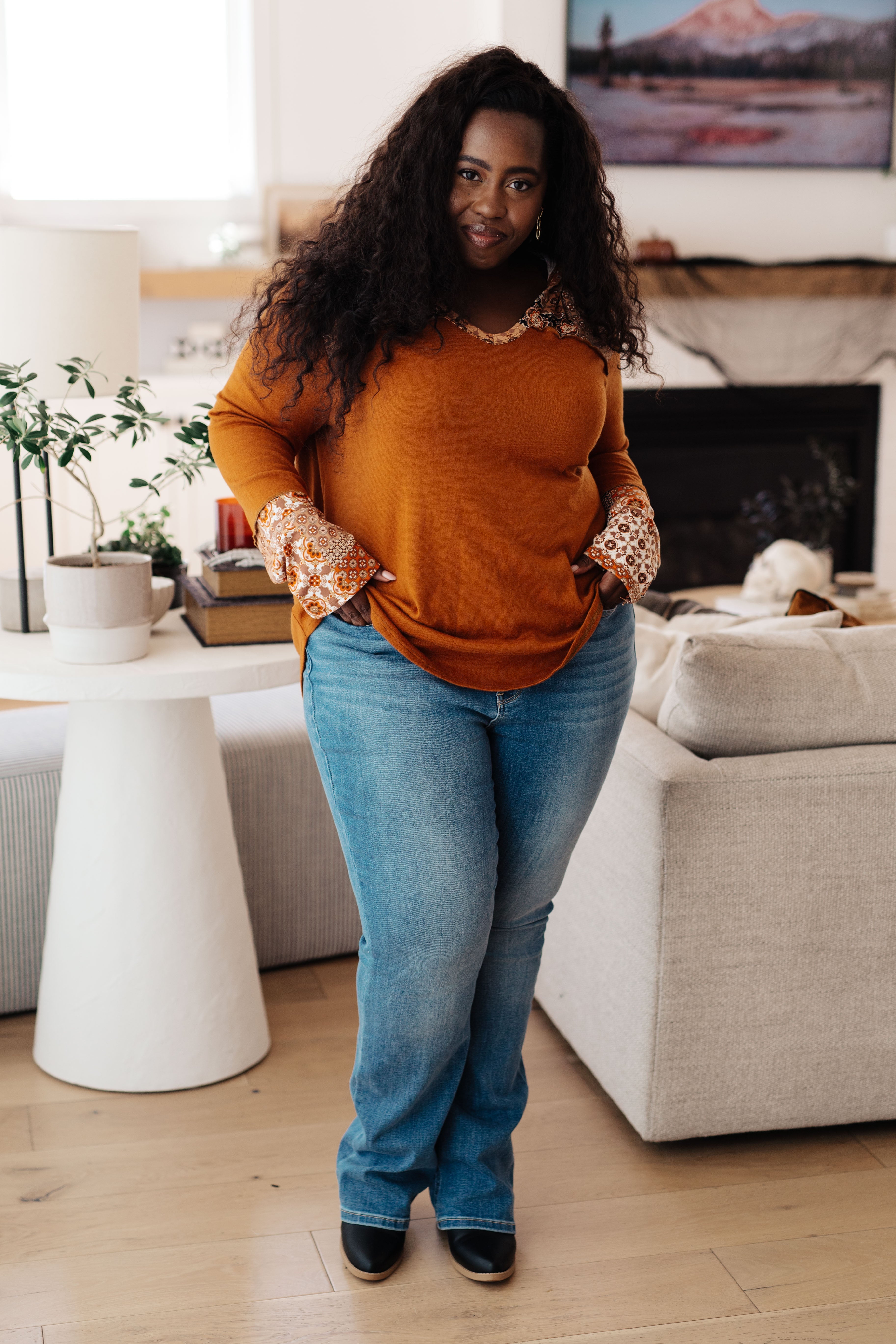 Plus-size woman wearing rust mixed print hoodie with relaxed fit paired with light blue jeans in cozy living room