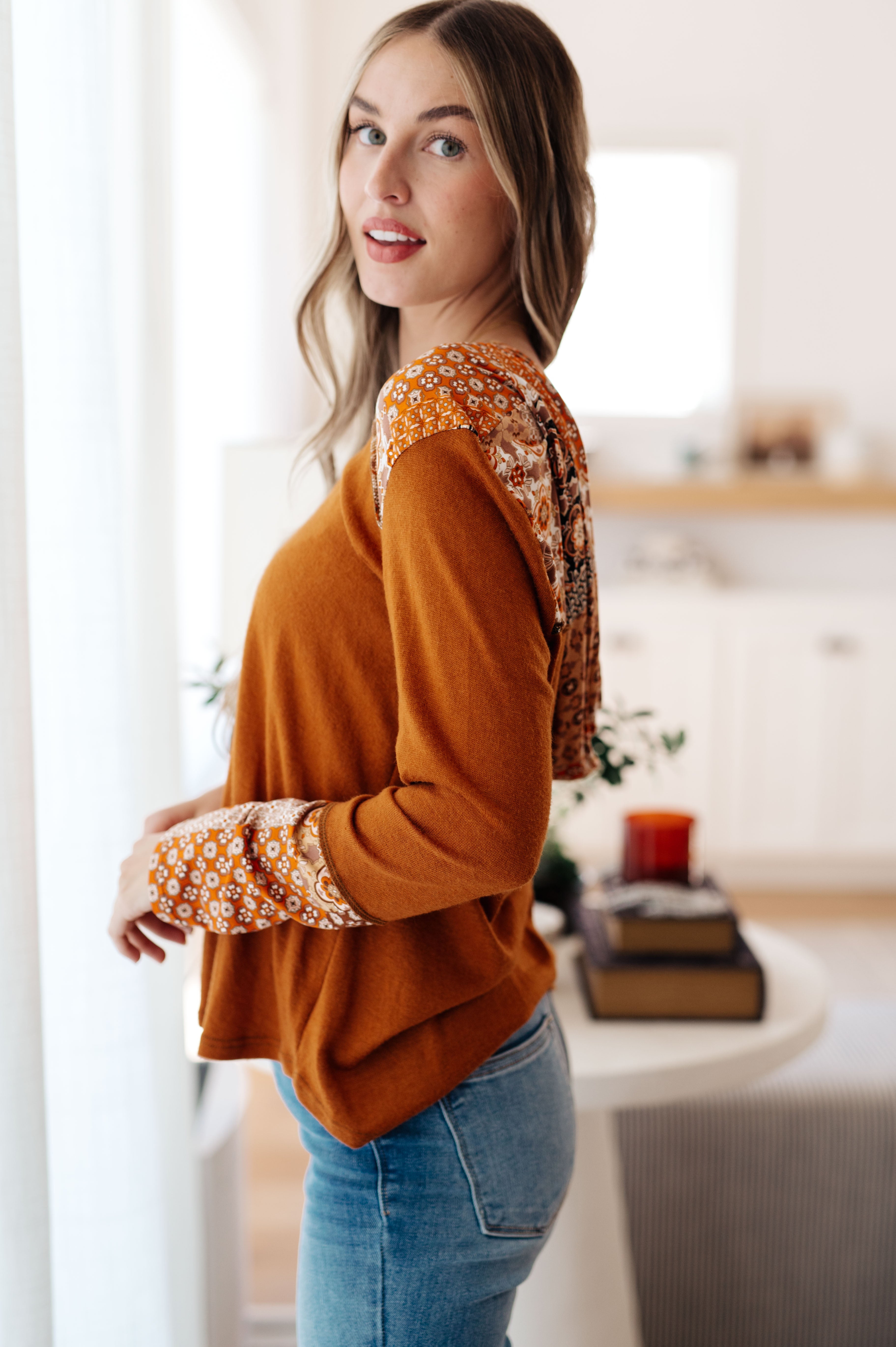 Woman wearing rust-colored mixed print hoodie with floral pattern on sleeves and hood in bright cozy room
