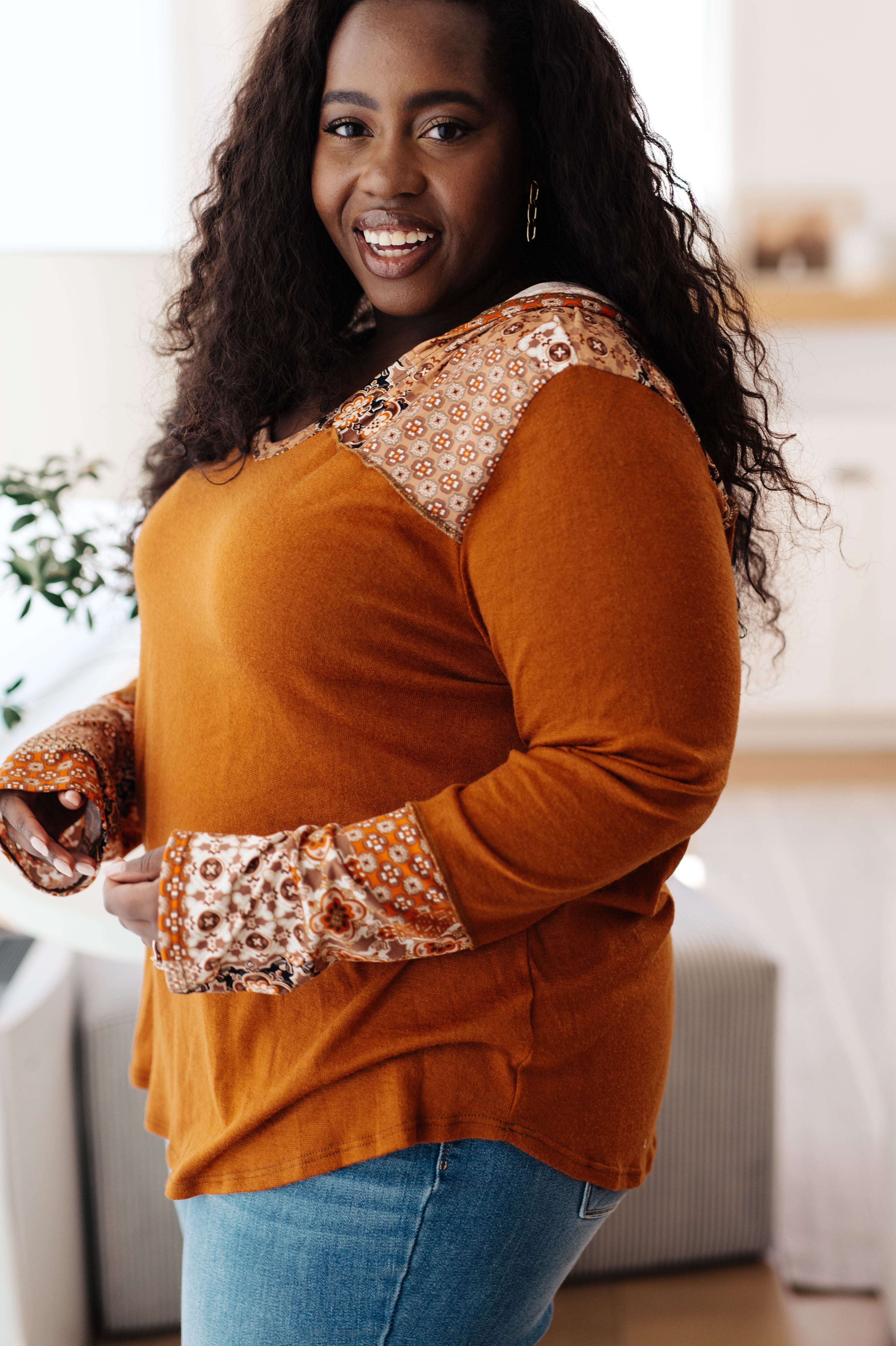 Smiling plus-size woman wearing rust-colored mixed print hoodie with floral accents and blue jeans indoors