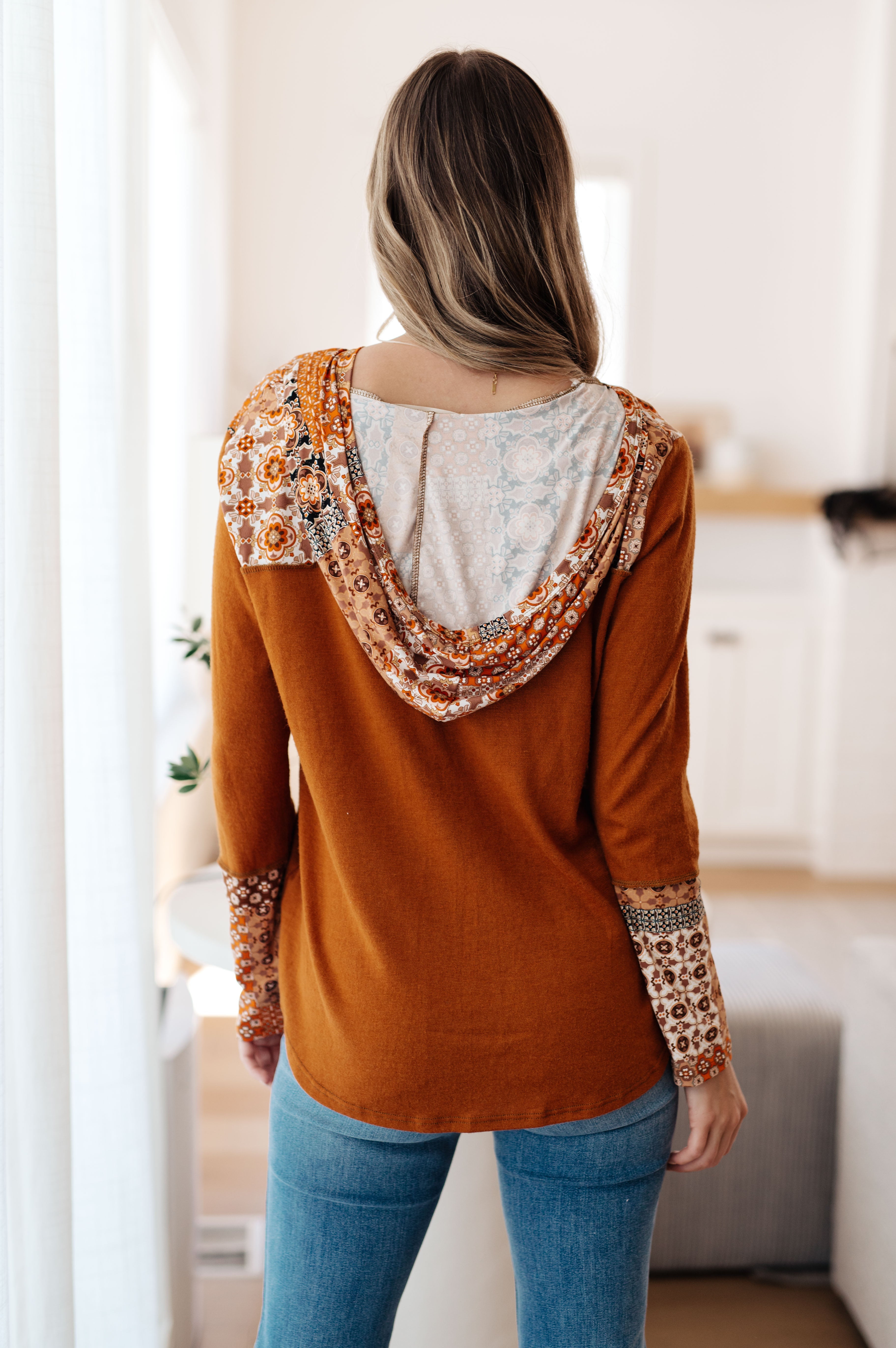 Back view of woman wearing rust-colored mixed print hoodie with patterned sleeves and hood in cozy indoor setting