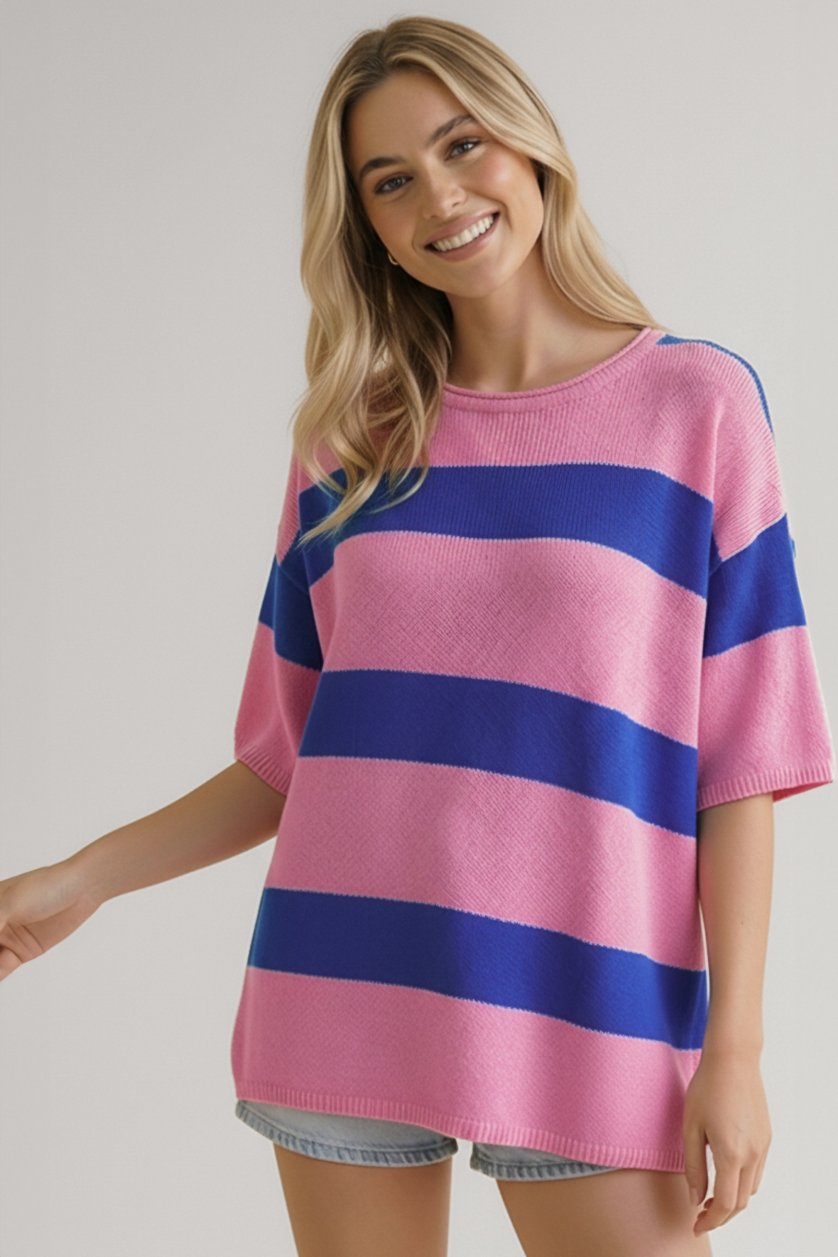 Smiling woman wearing a loose pink and blue wide-striped knit top with elbow-length sleeves and light denim shorts