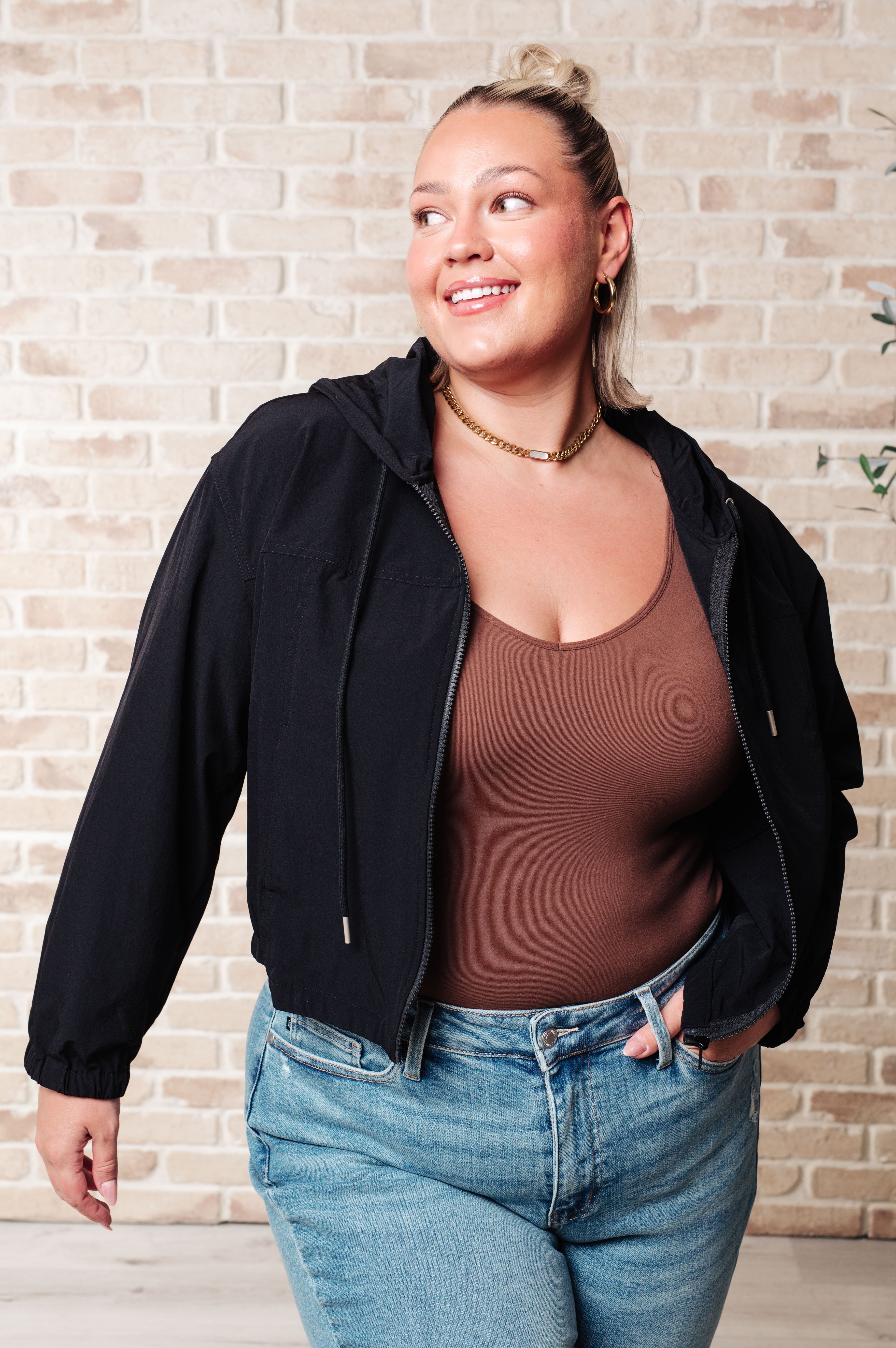 Plus-size woman wearing black zip-up hoodie over brown tank top paired with light blue jeans against brick wall background