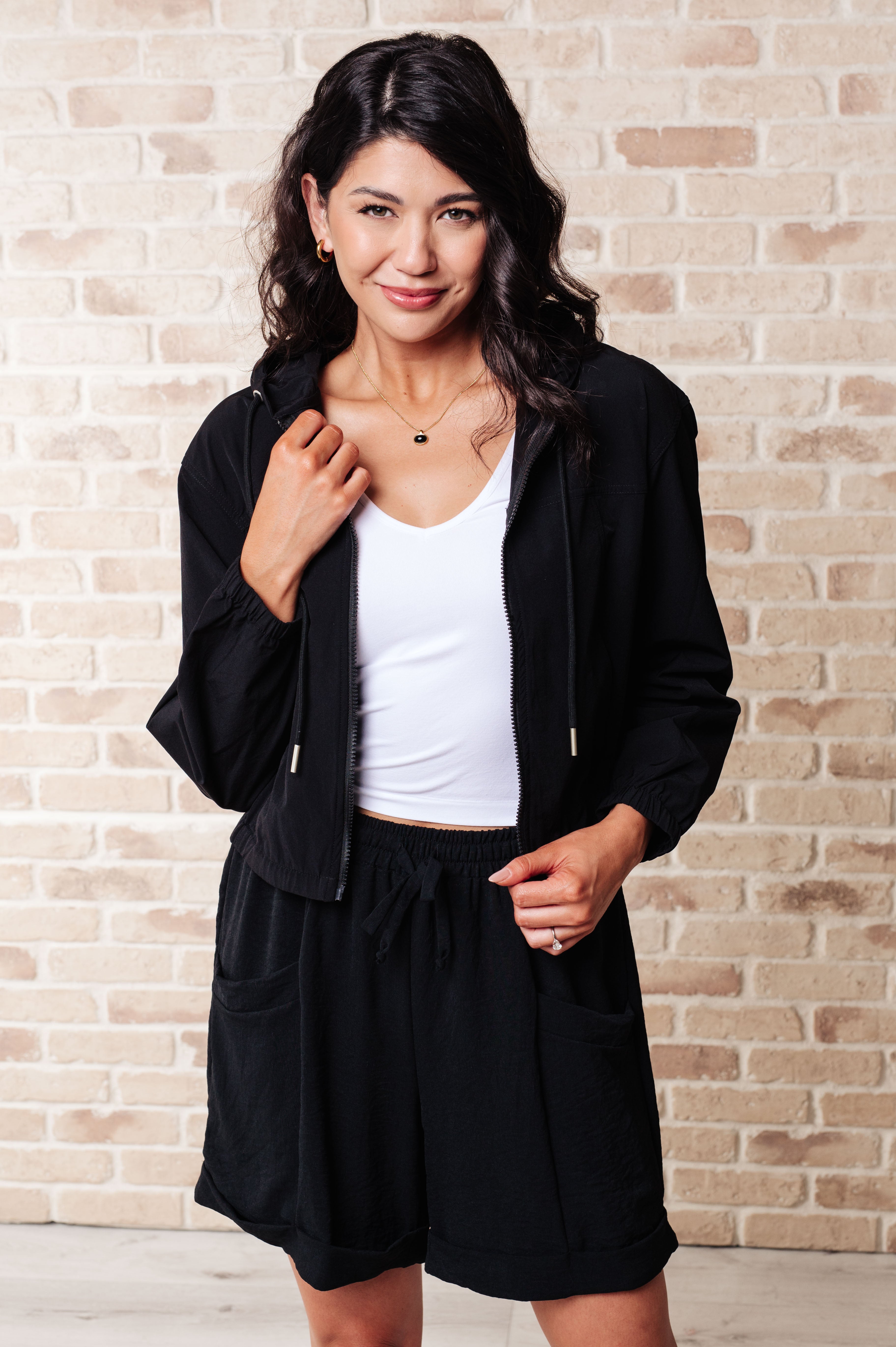 Woman wearing black zip-up jacket and matching shorts over white tank top, posing against light brick wall background