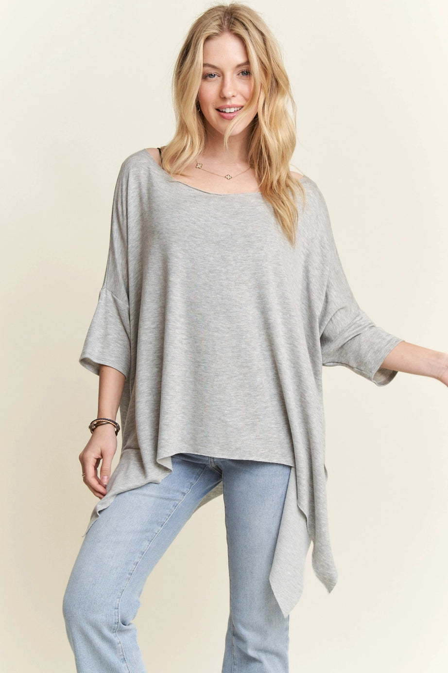 Woman wearing grey flowy top with raw edge shark-bite hem paired with light wash jeans and layered necklaces