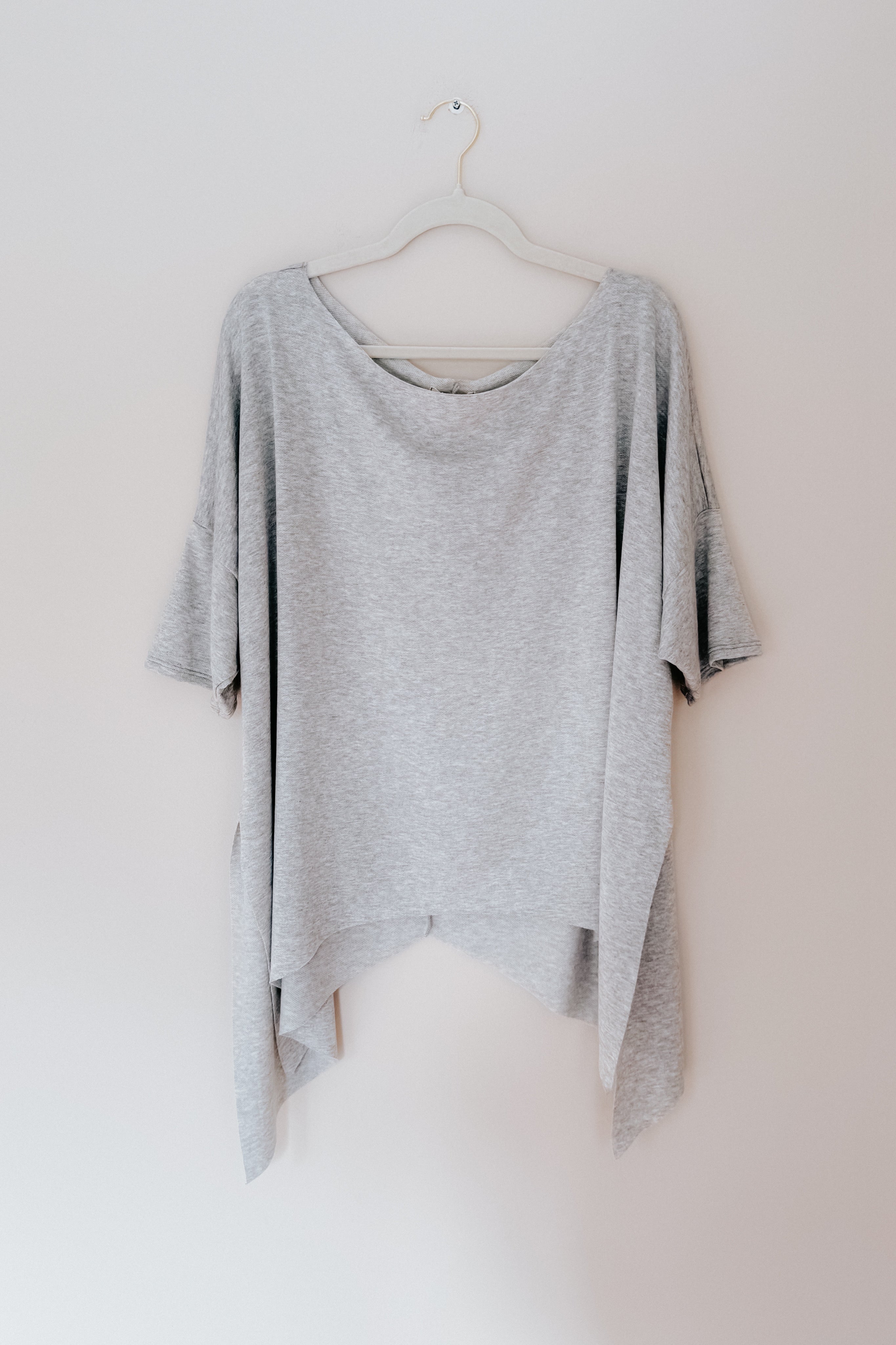 Grey flowy top with raw edge and shark-bite hem hanging on white hanger against plain wall background