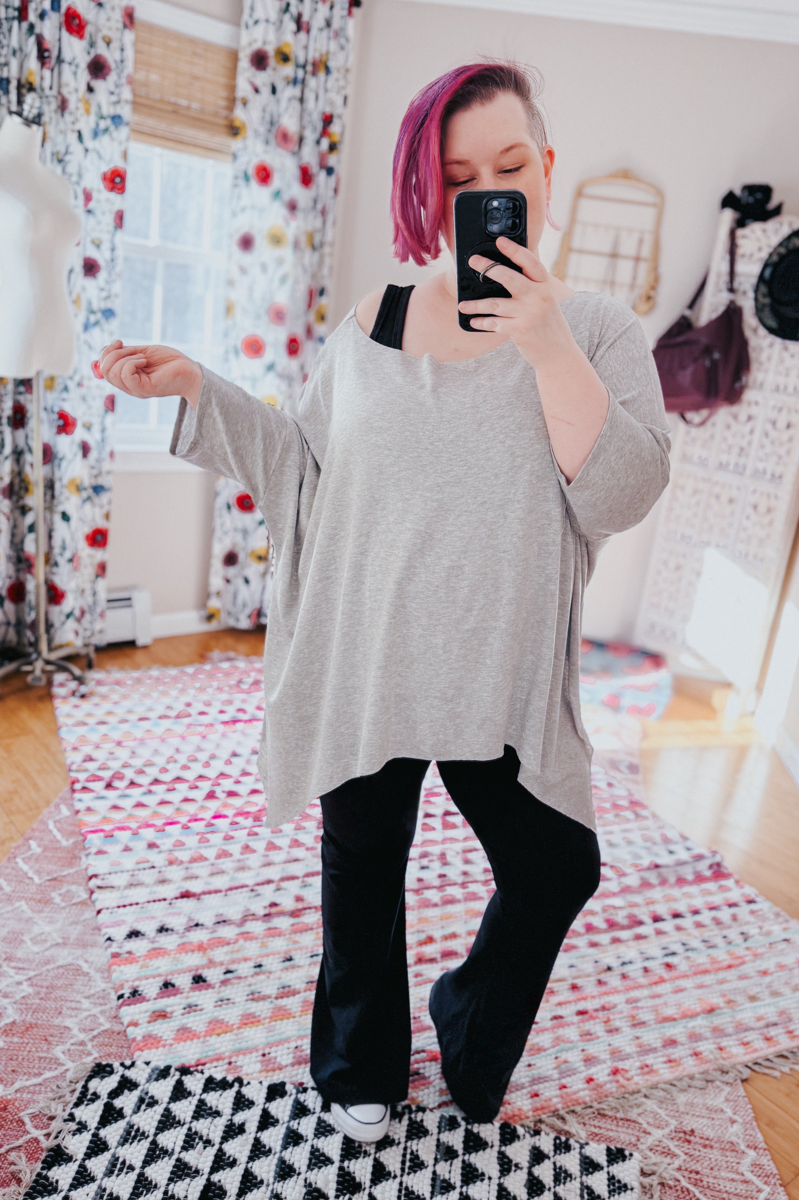 Woman with pink hair wearing a grey loose flowy top and black pants taking a selfie in a bright, patterned room