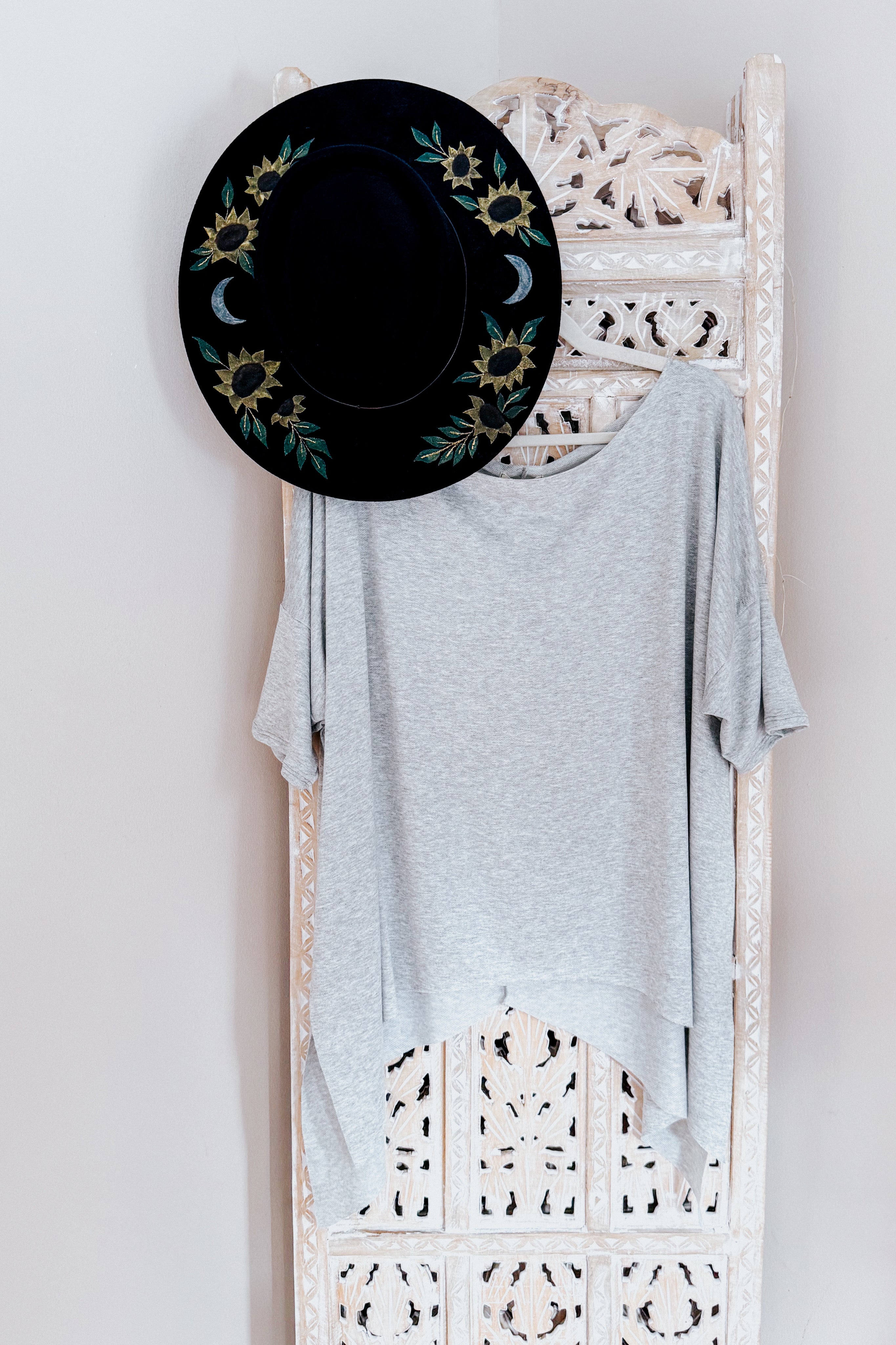 Light grey flowy top with raw edge shark-bite hem hanging on carved wooden panel with black floral hat above