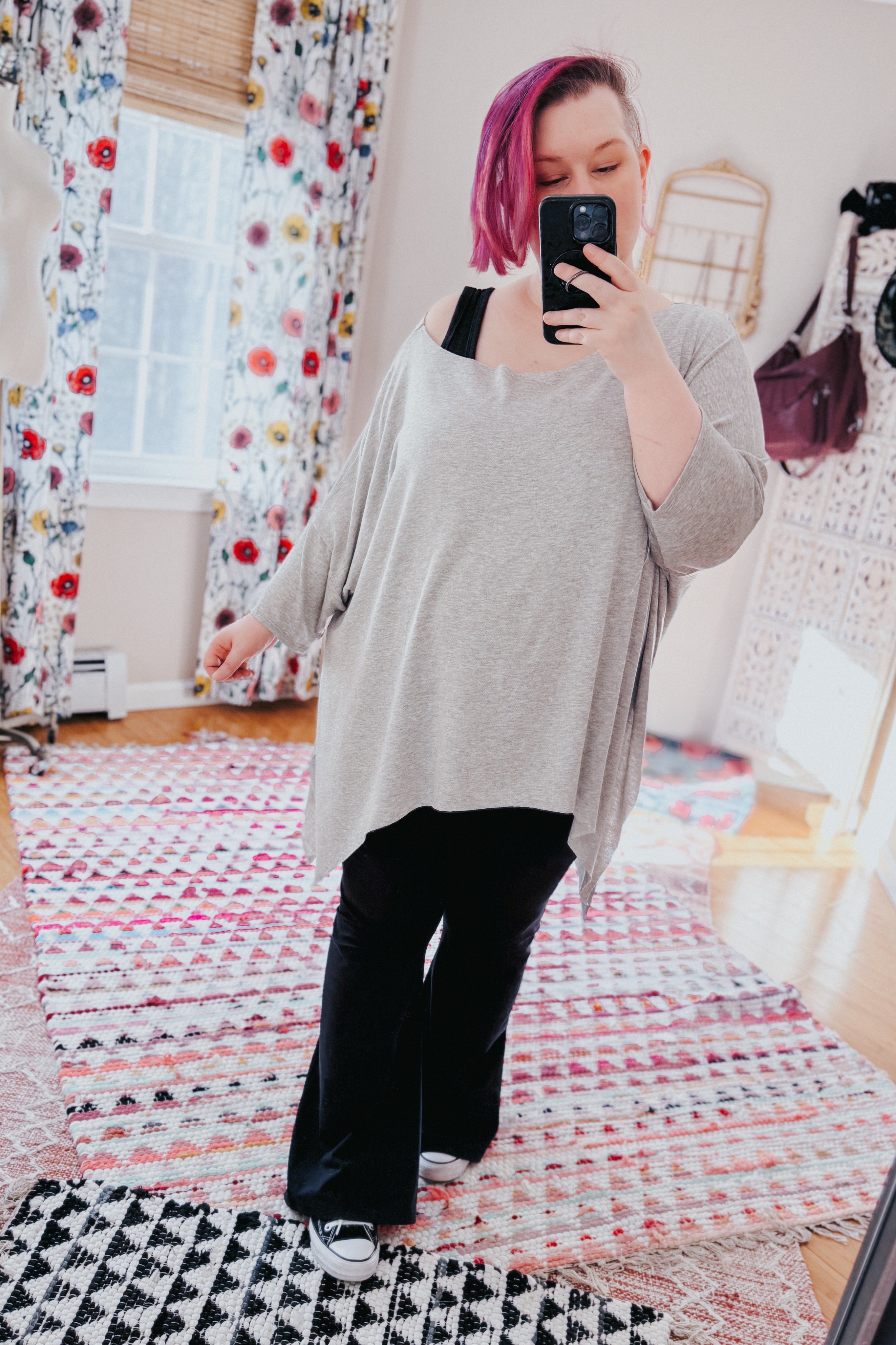 Woman wearing loose grey flowy top with raw edges over black pants taking mirror selfie indoors