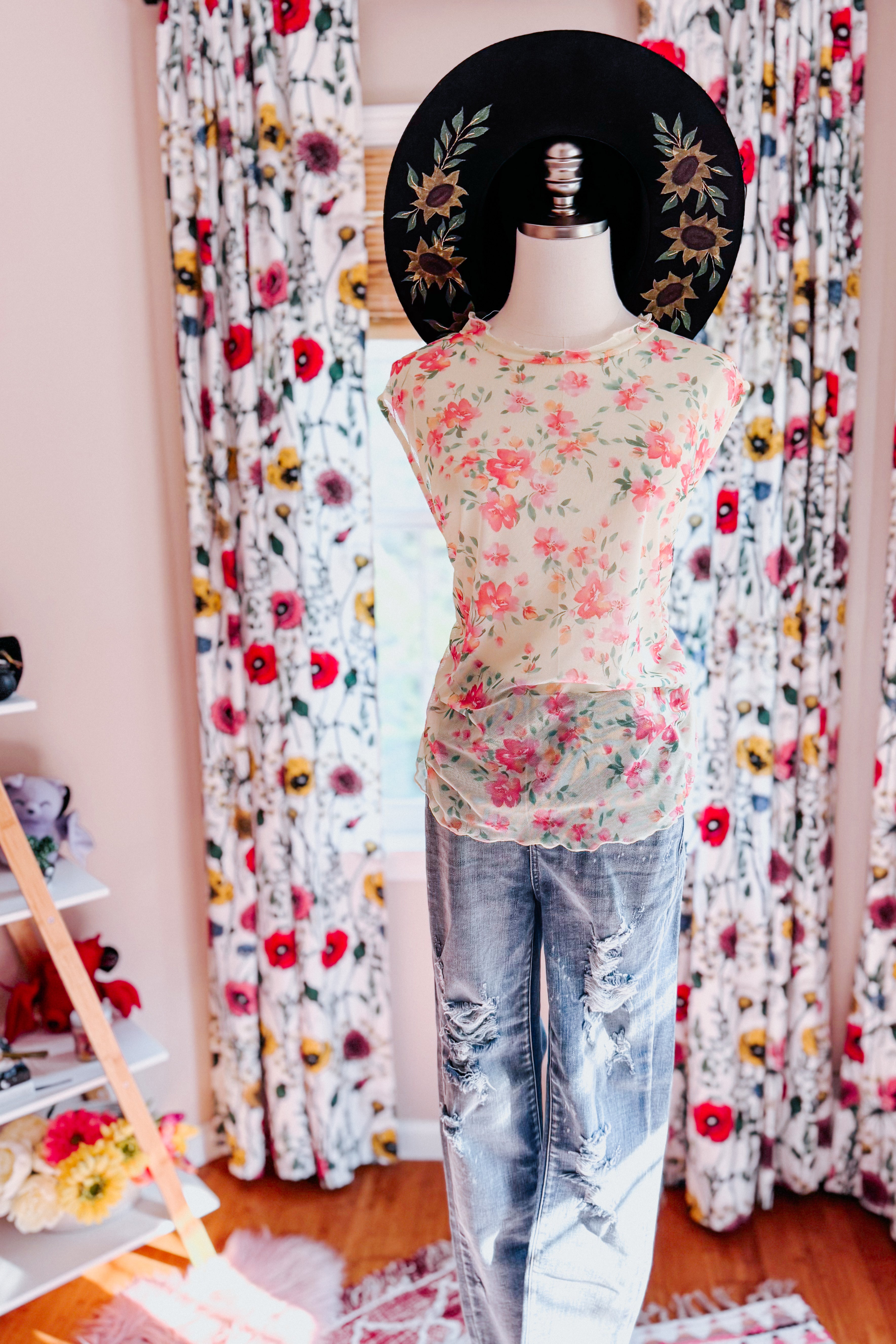Mannequin dressed in sheer floral top layered over distressed blue jeans with large black sunhat decorated with sunflowers