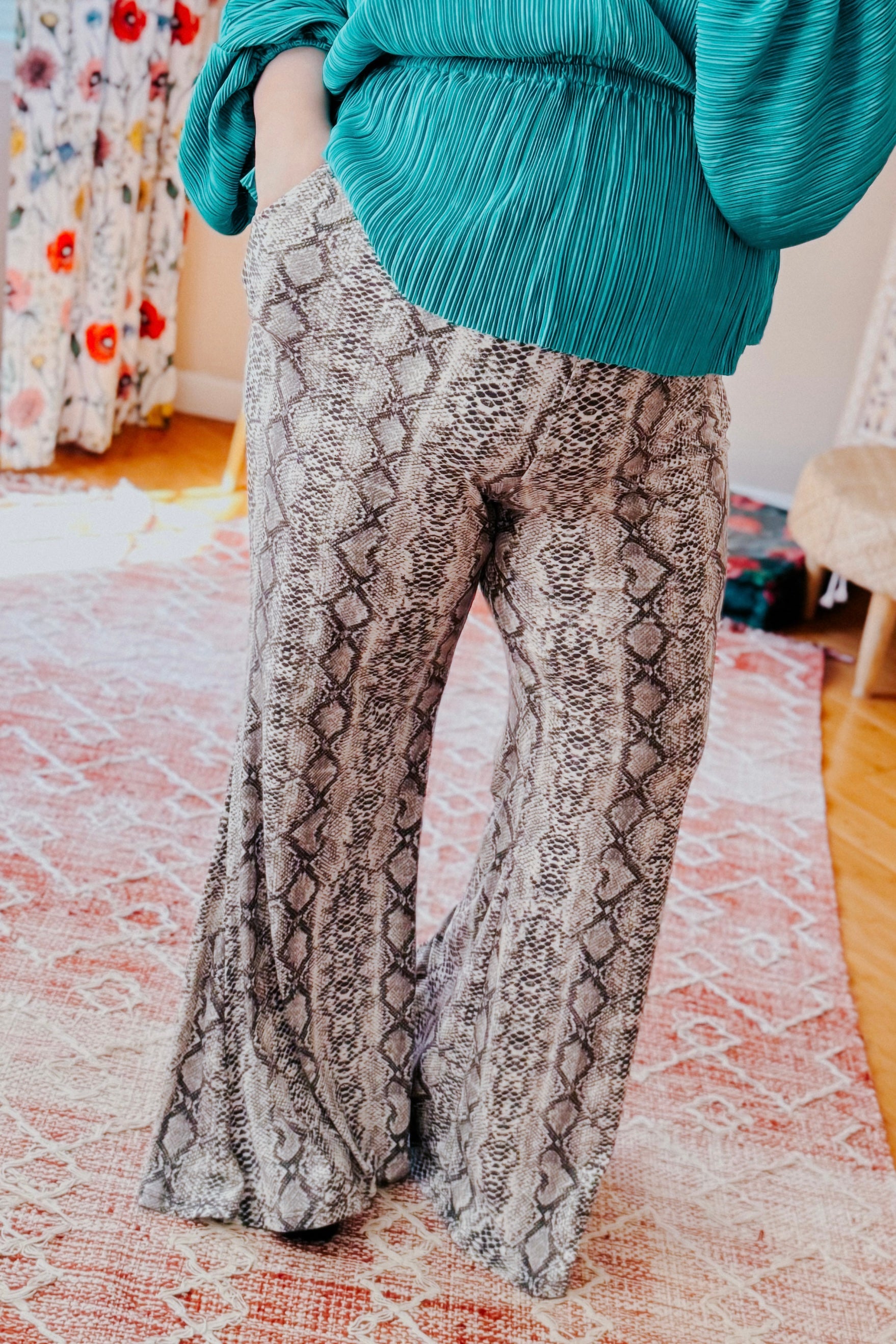 Woman wearing grey snake print flare pants paired with pleated teal top standing on patterned rug indoors