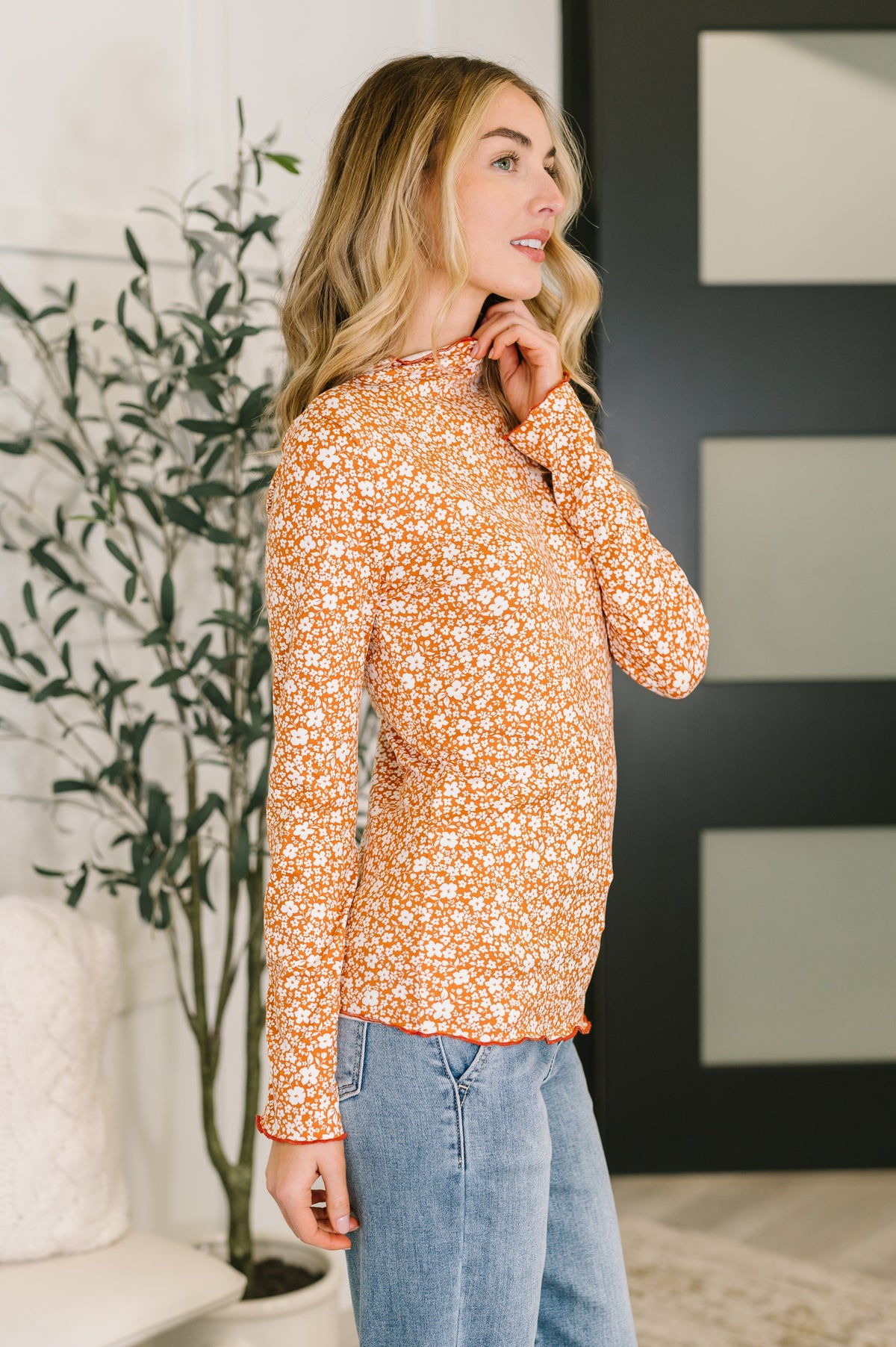 Side profile of a woman wearing an orange floral long sleeve top with lettuce trim and light wash jeans near a leafy houseplant