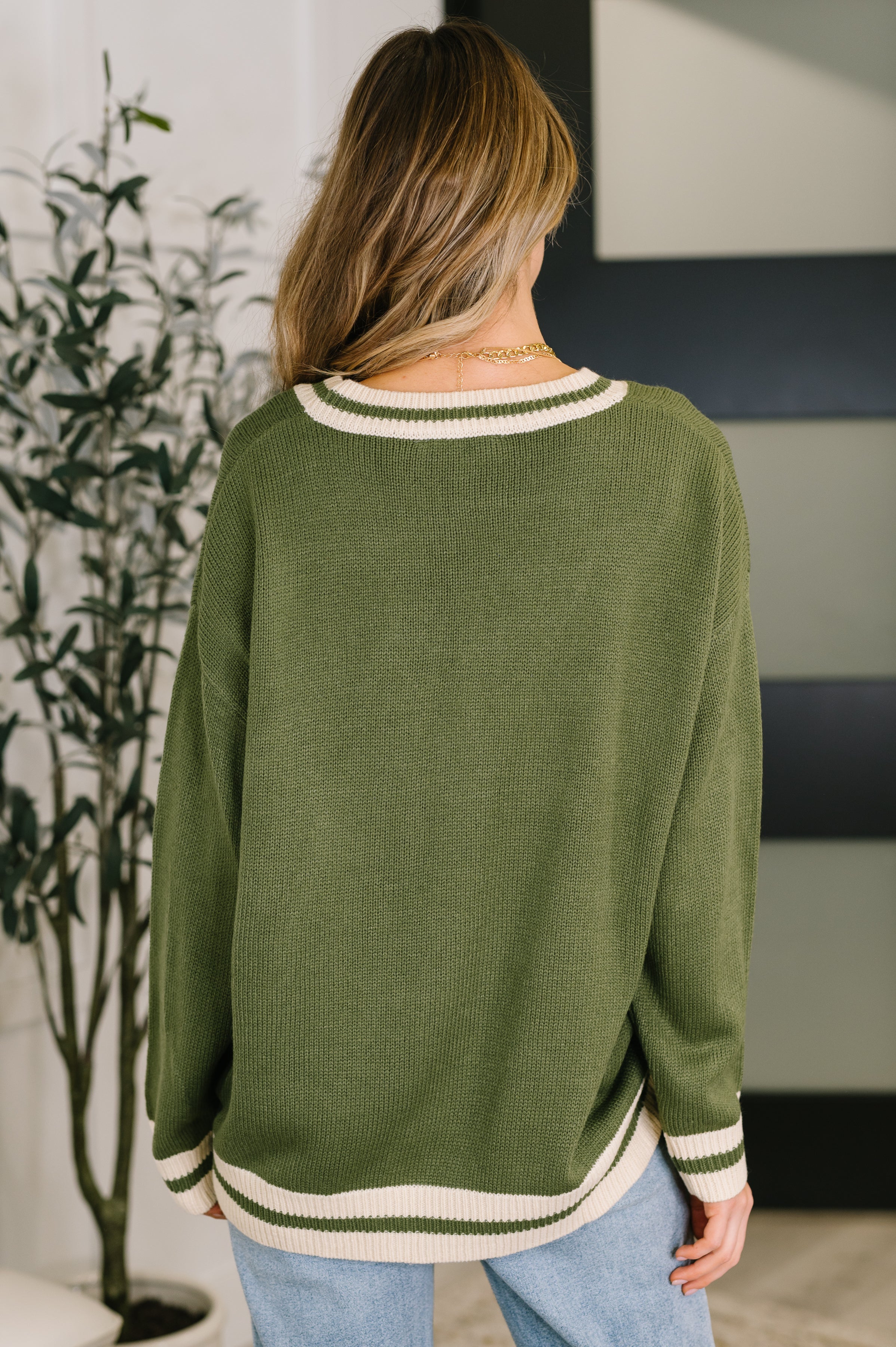 Woman with her back facing the camera wearing a green cardigan with white trim and blue jeans in an indoor setting