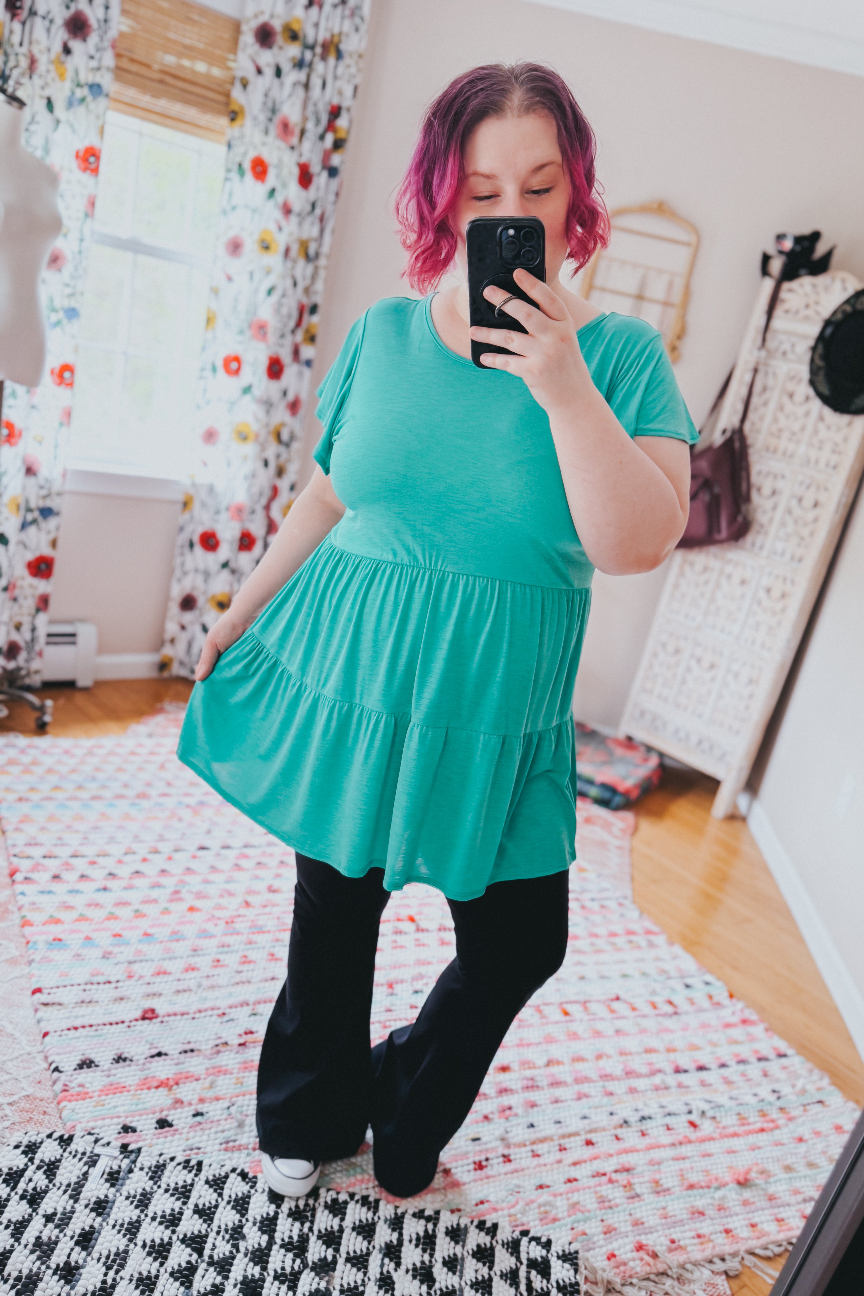 Woman with pink hair wearing teal babydoll top and black pants taking mirror selfie indoors on patterned rugs