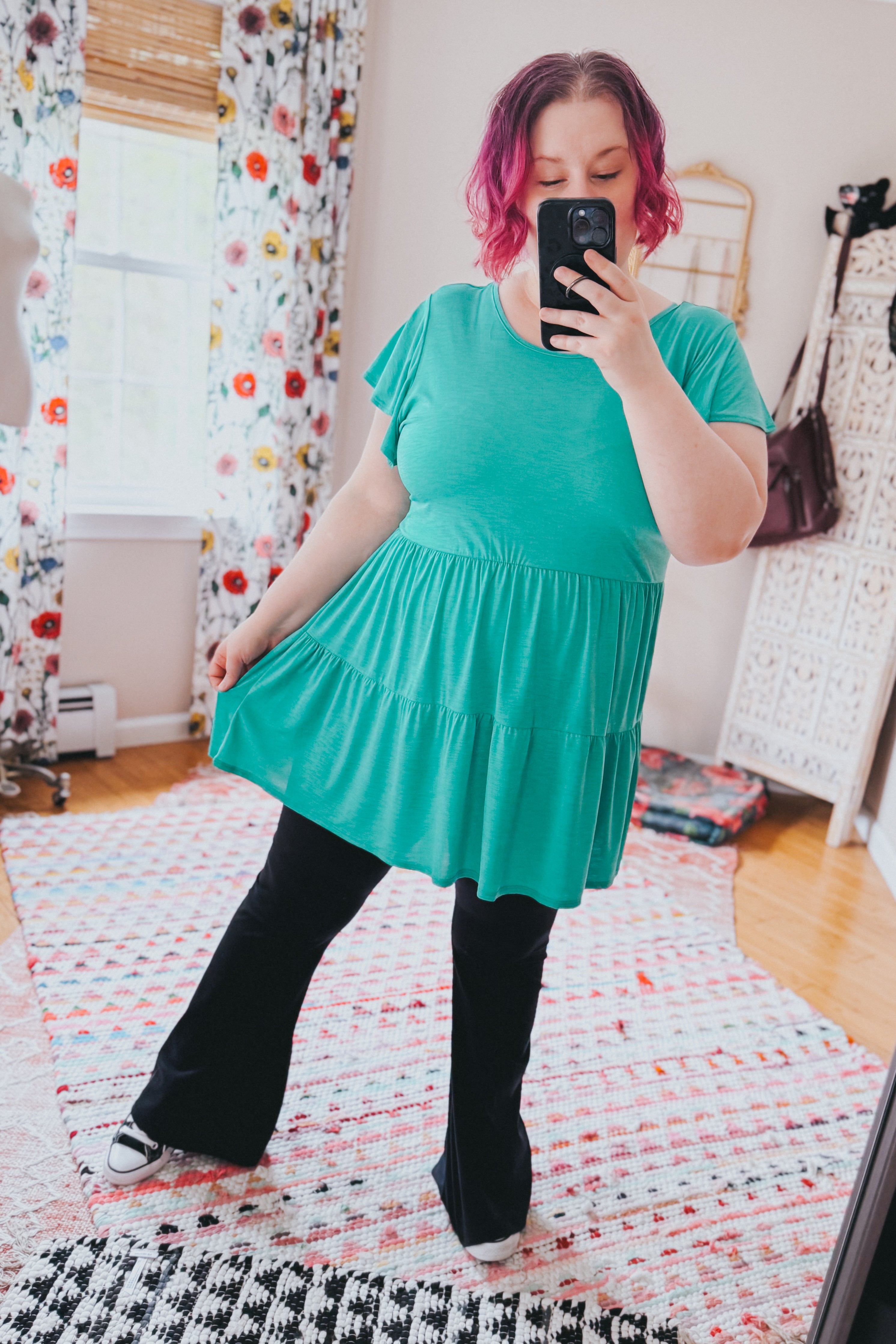 Woman with pink hair wearing a teal babydoll top paired with black pants taking a mirror selfie in a bright room