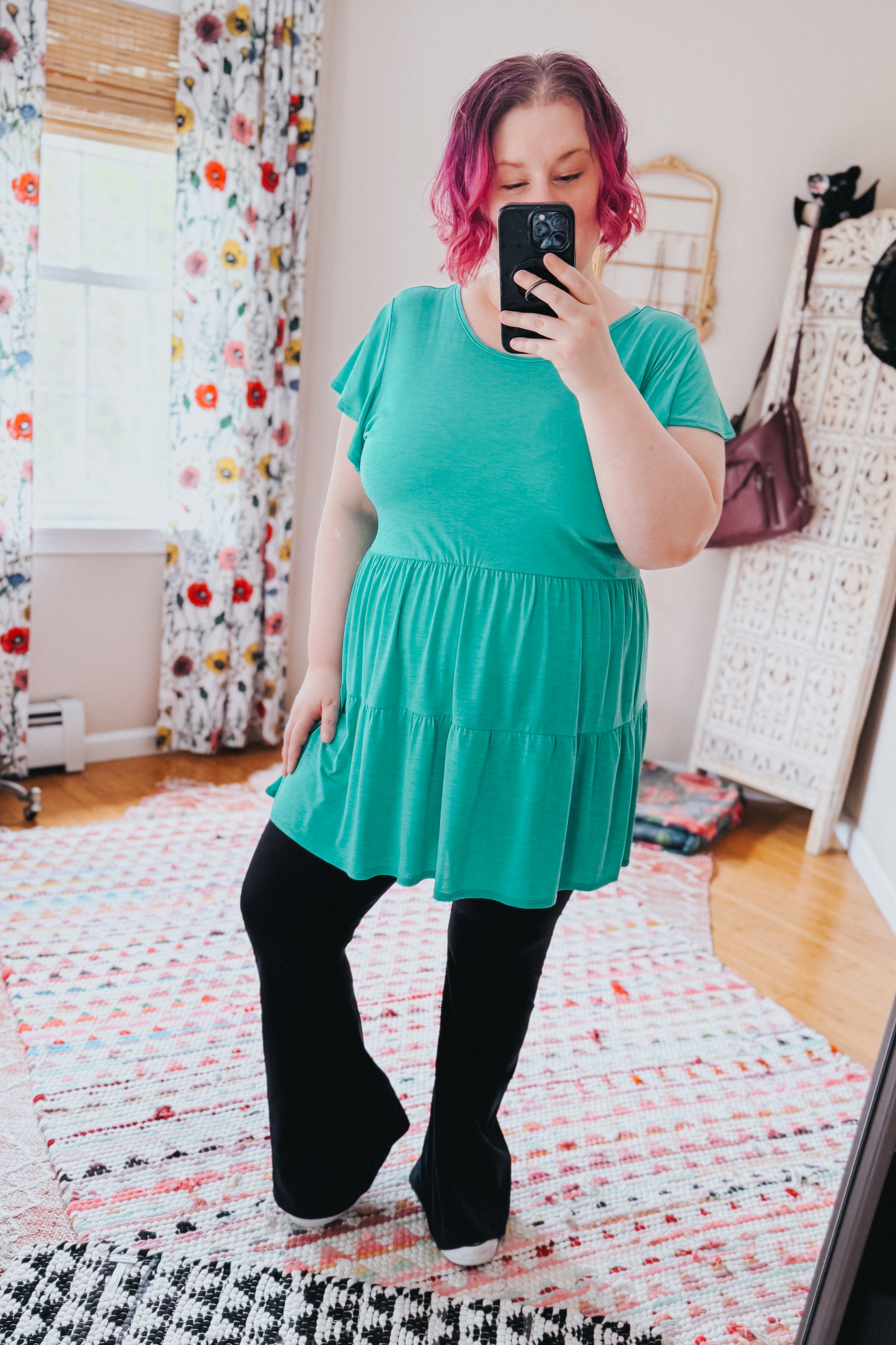 Woman with pink hair wearing teal babydoll top and black pants taking mirror selfie in cozy room with patterned rugs