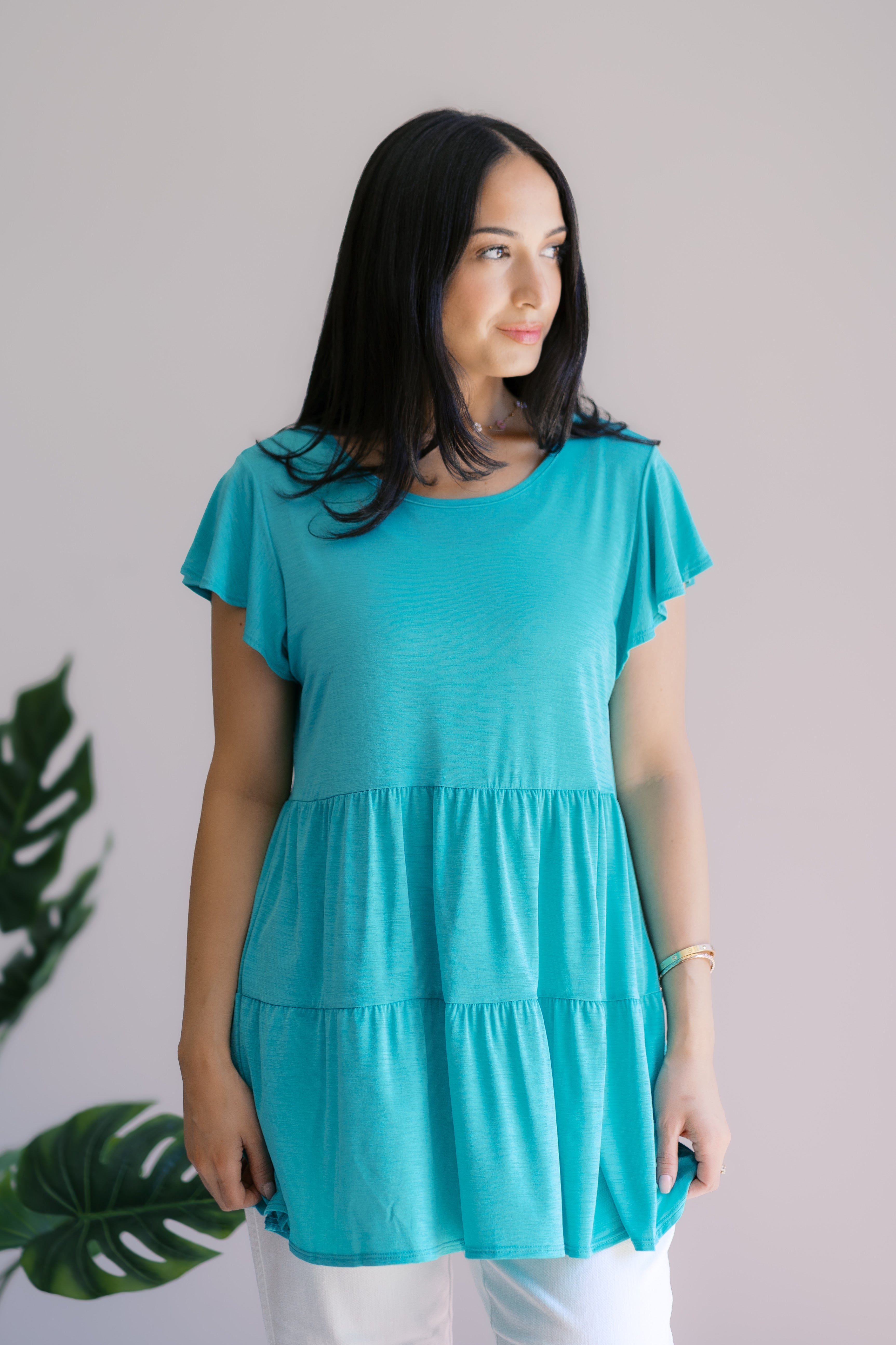 Woman wearing teal babydoll top with short flutter sleeves standing indoors near green plant leaves