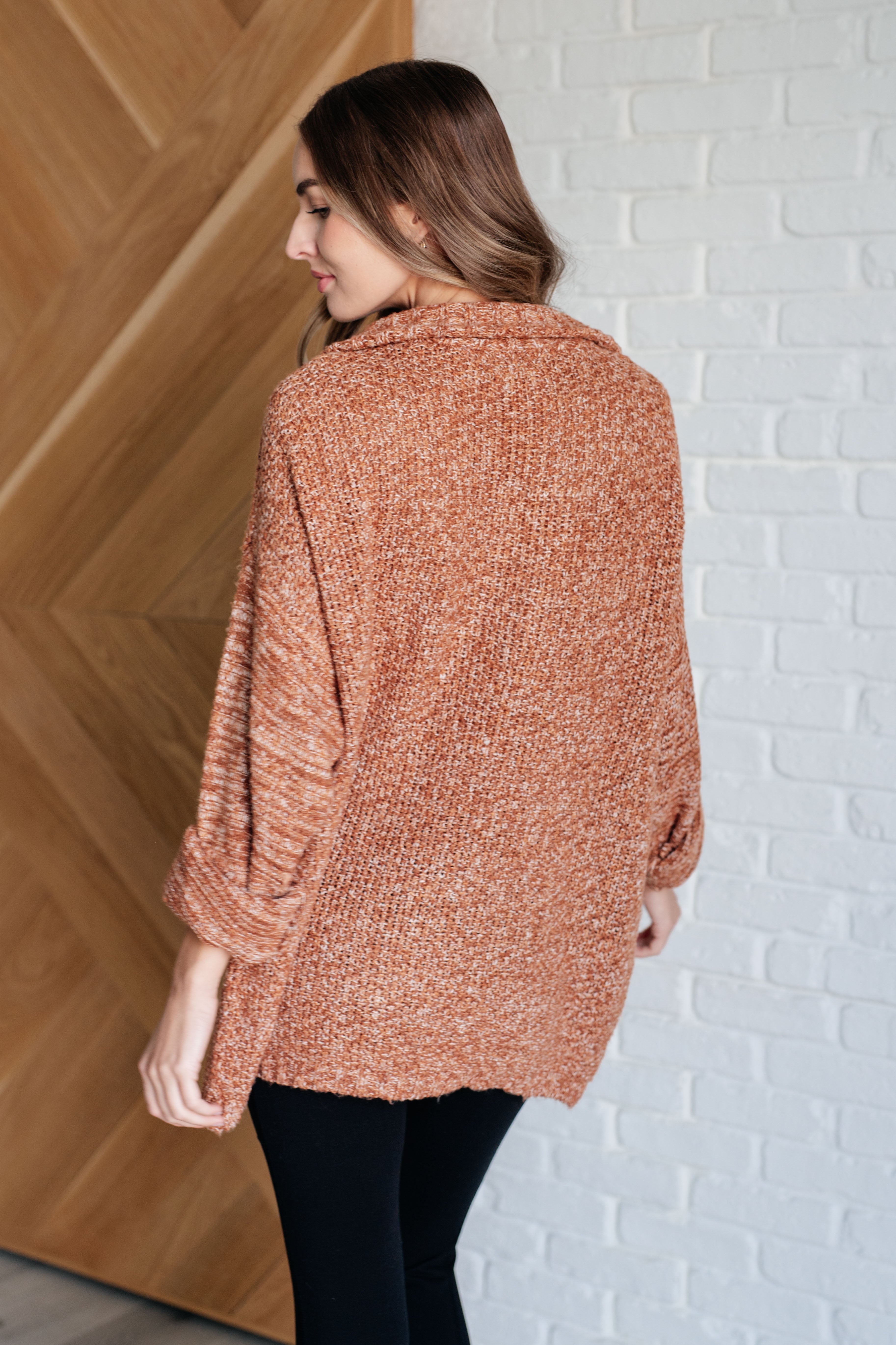 Woman wearing a rust colored oversized long-sleeve sweater and black leggings standing against a white brick wall.