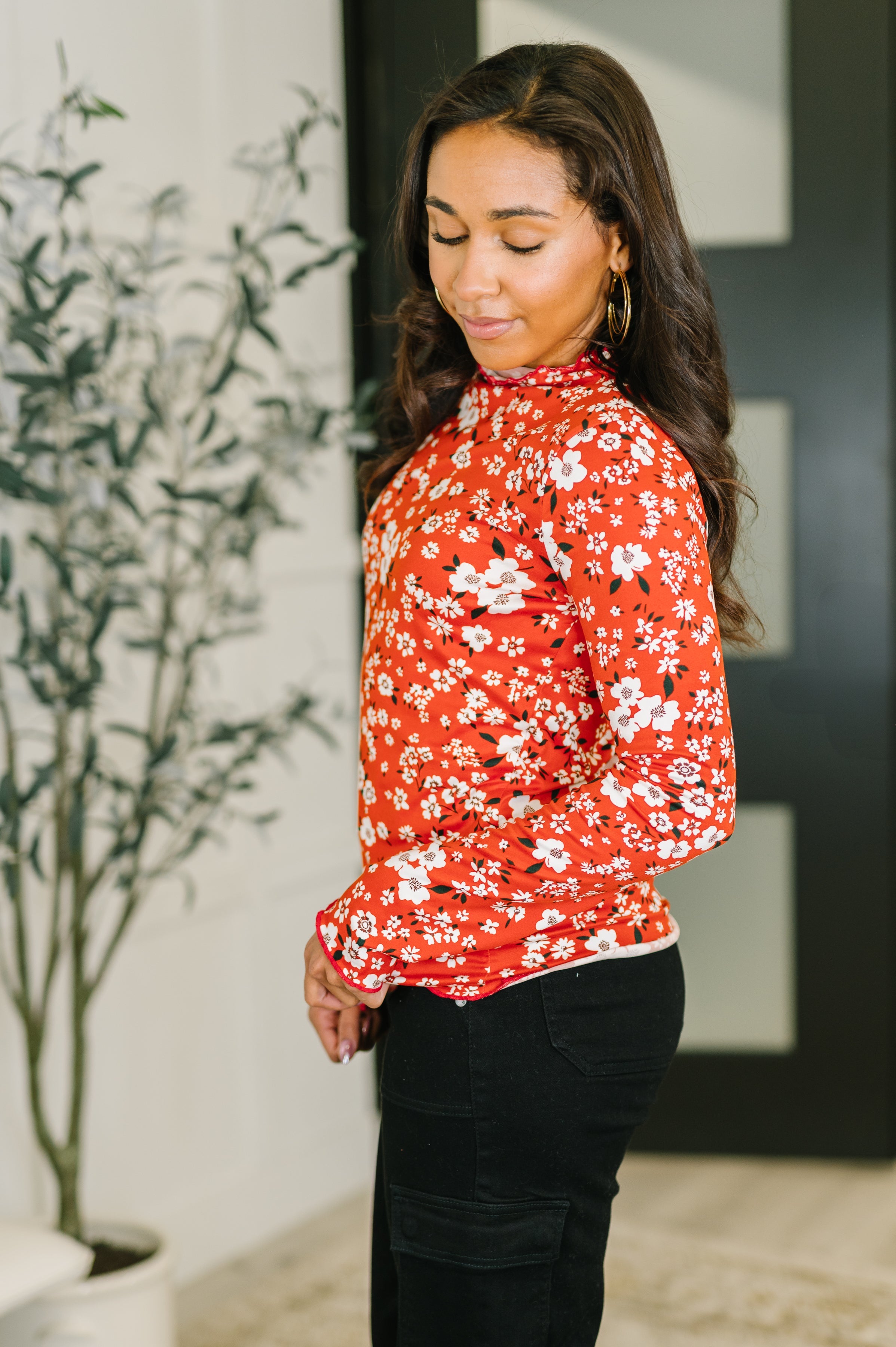 Woman wearing a vermillion long-sleeve top with white floral print and black pants standing indoors facing sideways