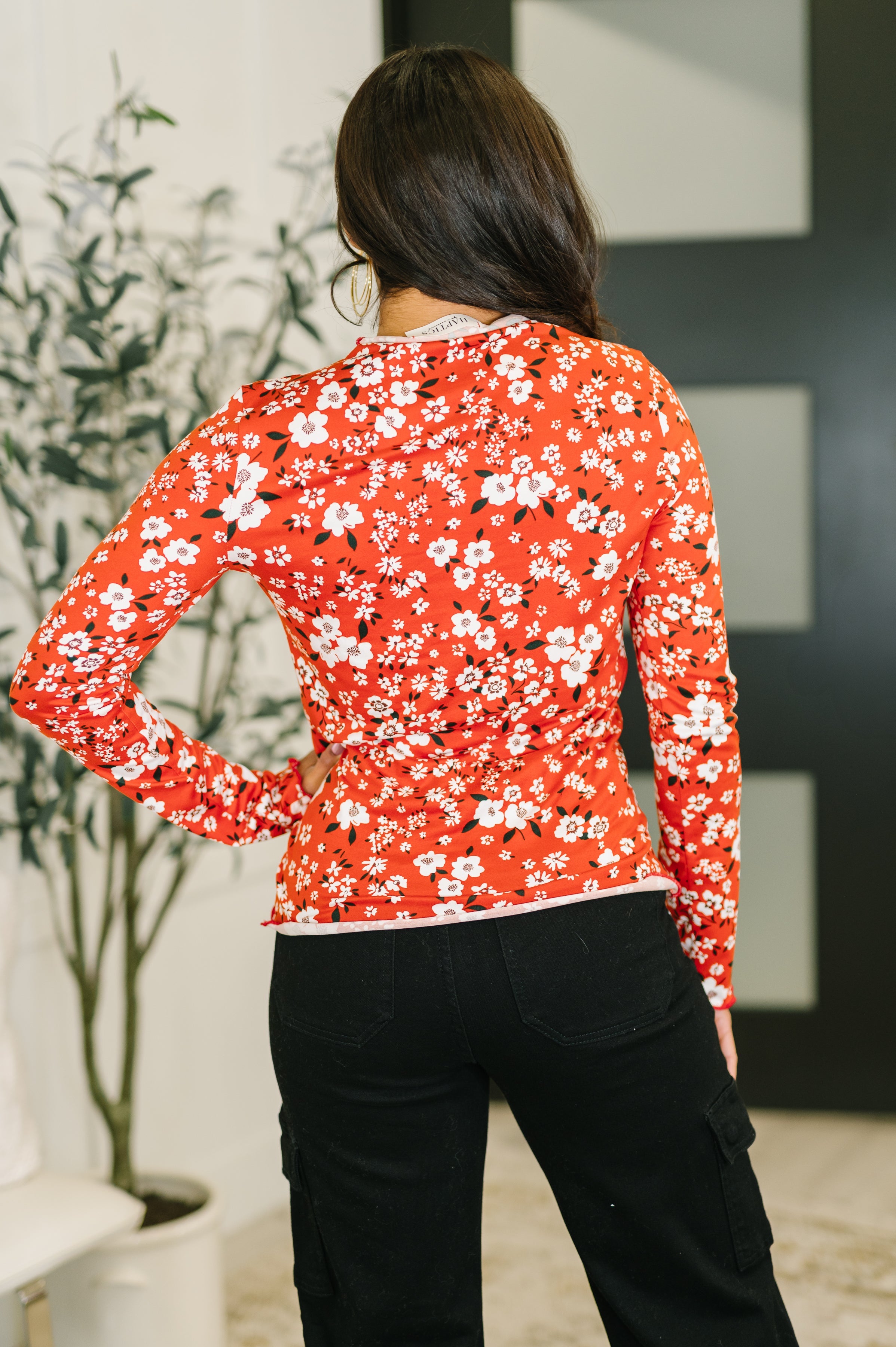 Woman wearing a vermillion long sleeve top with white floral print and black pants standing indoors with hand on hip
