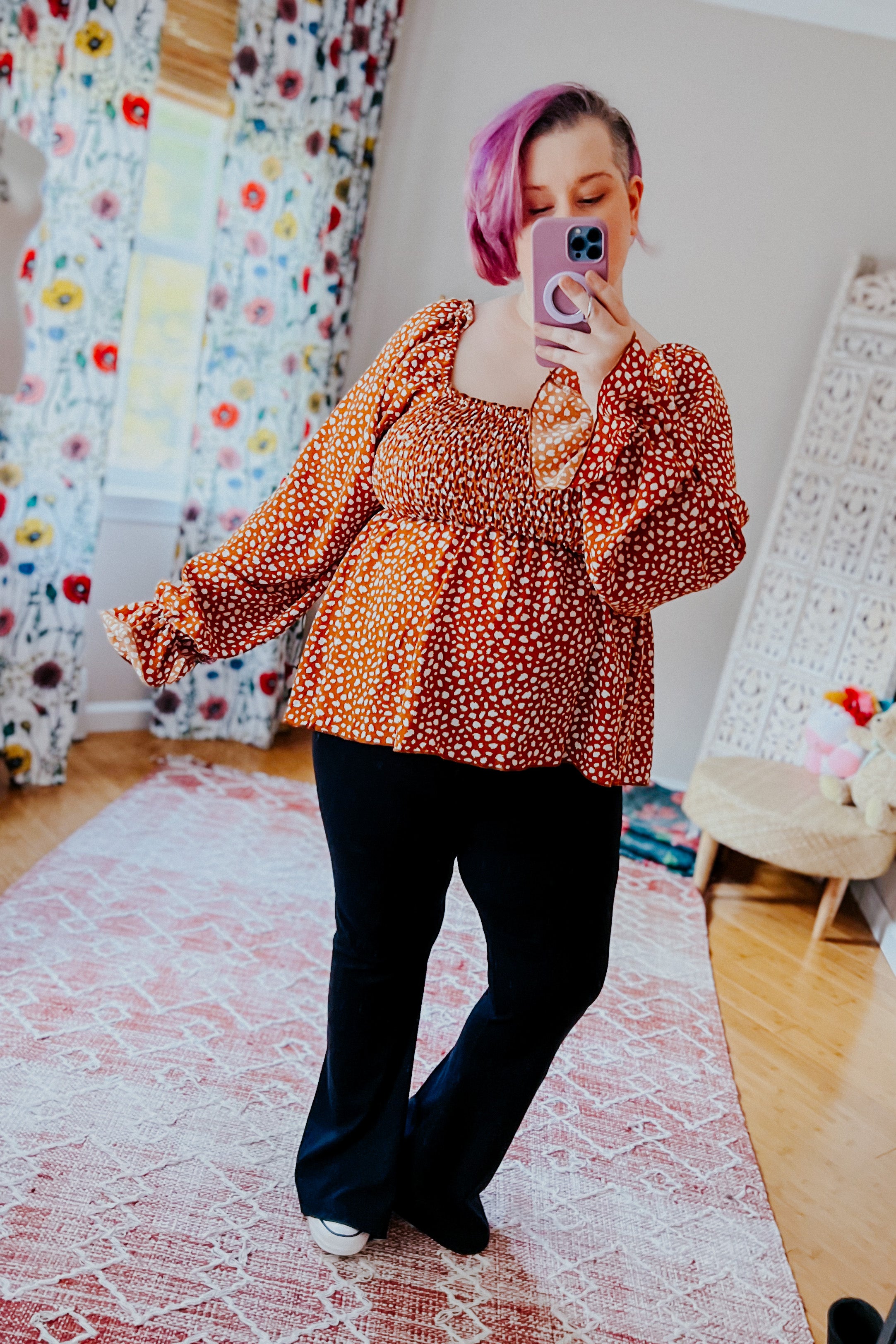 Woman with purple hair wearing rust smocked top with ruffle hem and black pants taking selfie indoors in bright room