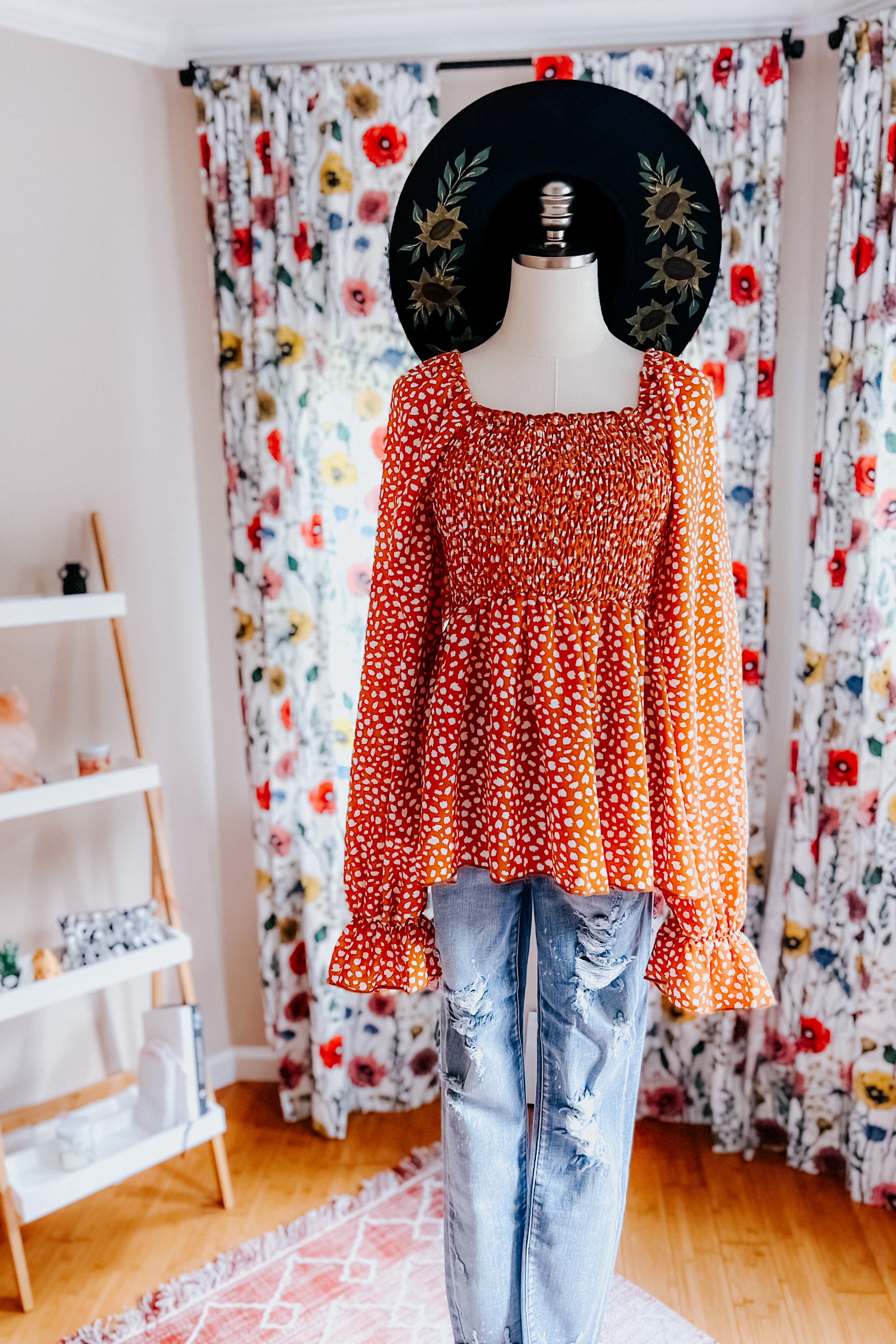 Rust-colored smocked top with ruffle hem and long puffed sleeves styled with distressed light-wash jeans on mannequin