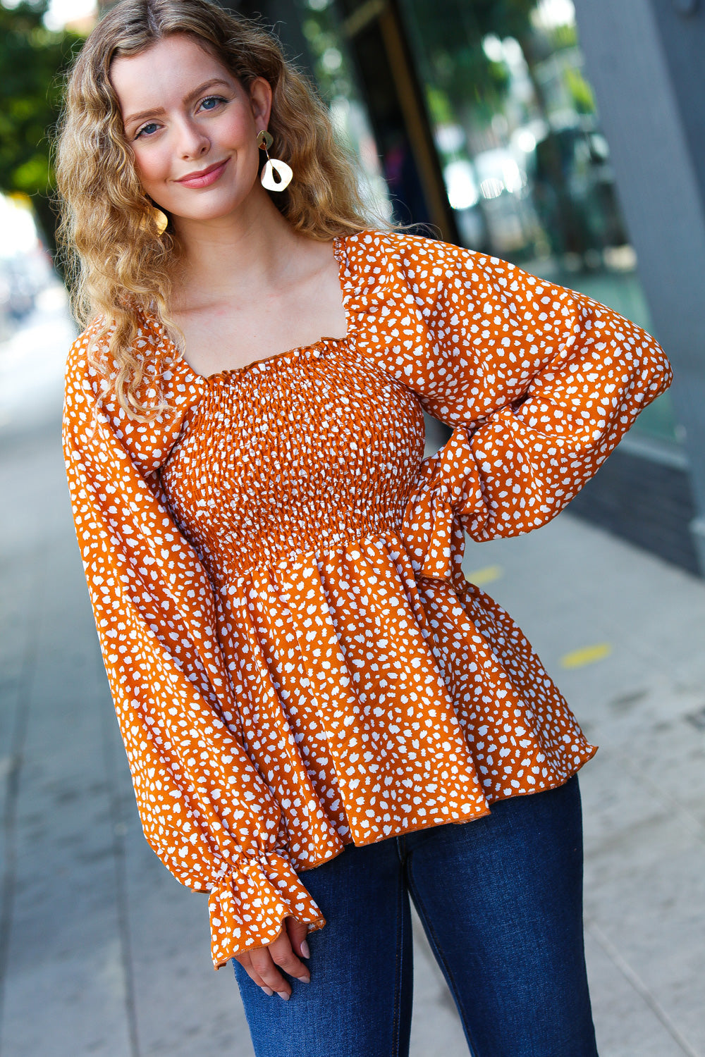 Woman wearing rust smocked top with ruffle hem and long puff sleeves standing outdoors on sidewalk