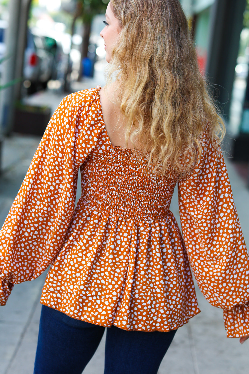 Woman with wavy hair wearing rust-colored smocked top with ruffle hem and spotted pattern, seen from behind outdoors