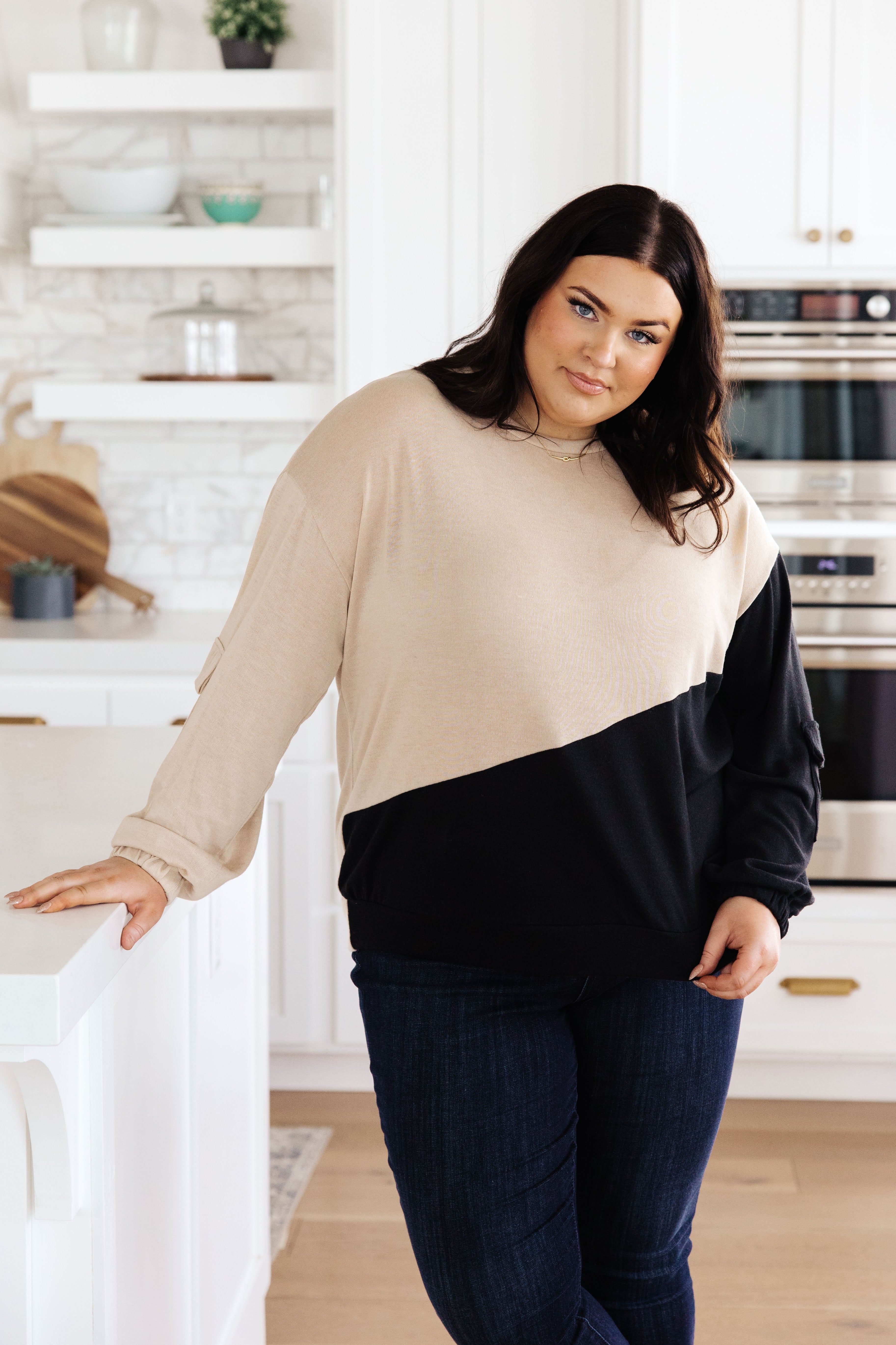 Woman wearing tan and black color block top with cargo sleeves posing in modern kitchen with white cabinets