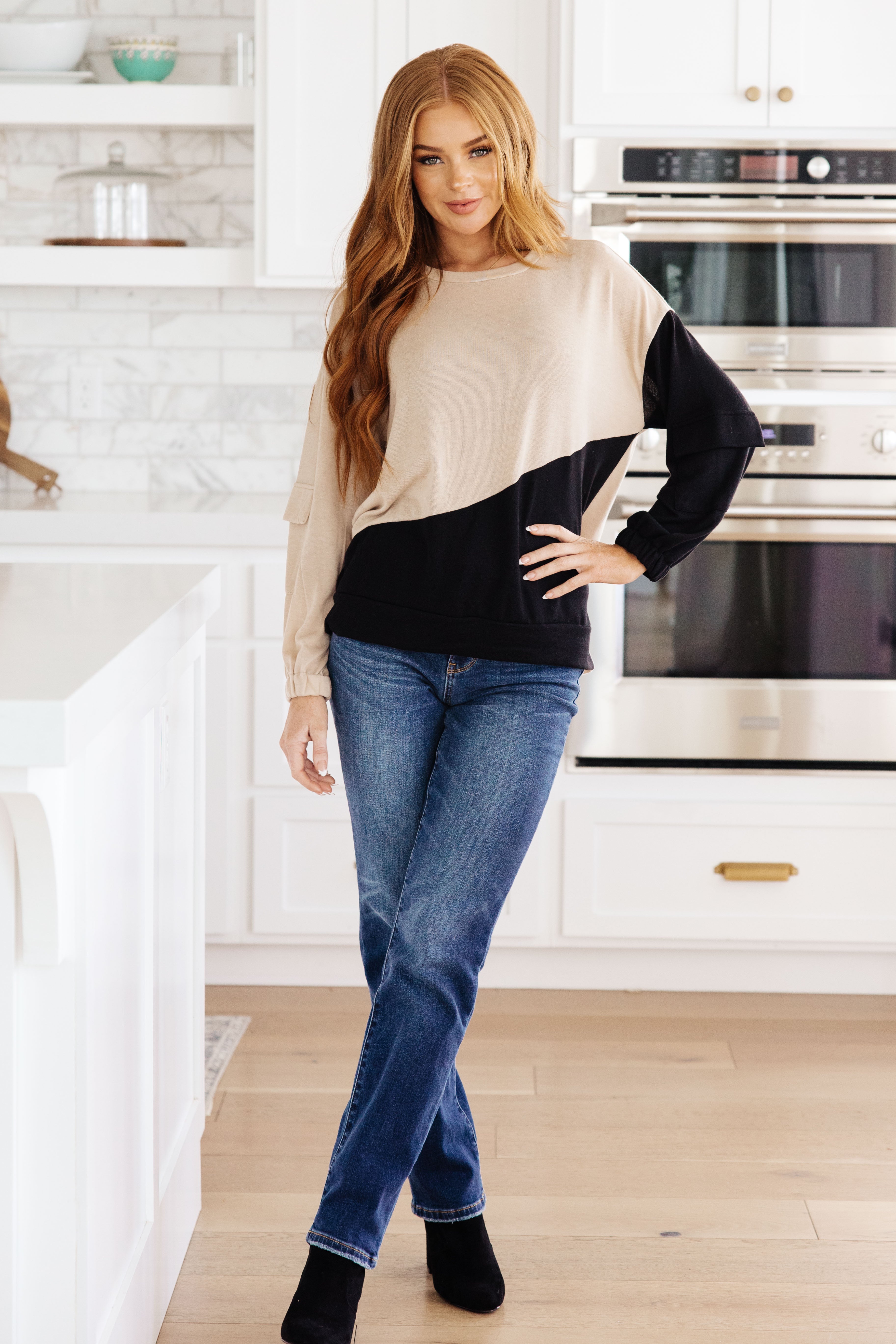 Woman wearing tan and black color block top with cargo sleeves paired with blue jeans and black boots indoors