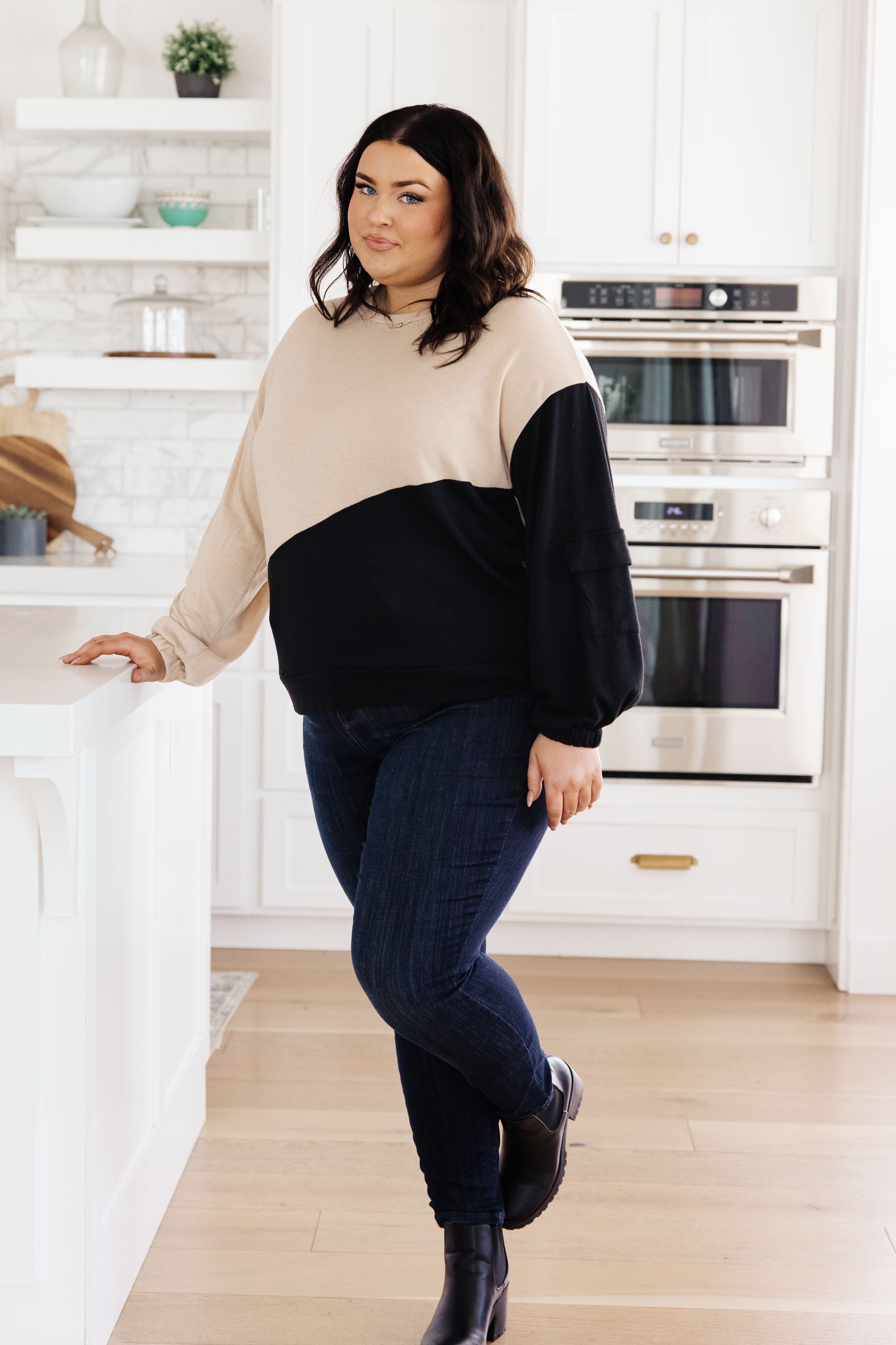 Woman wearing tan and black color block top with cargo sleeve detail paired with dark jeans in bright kitchen setting