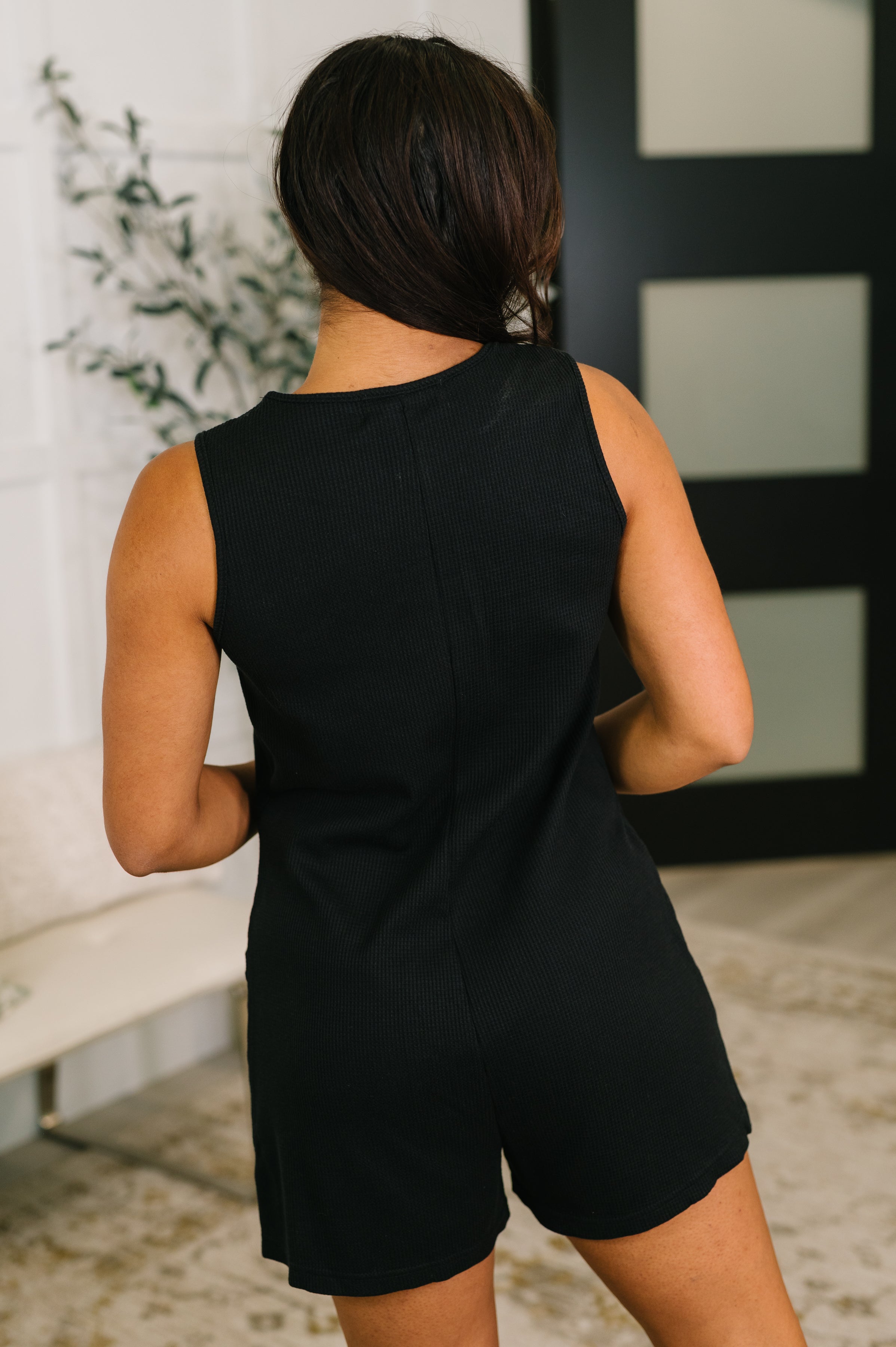 Woman wearing a black sleeveless romper with subtle ribbed texture in a bright, modern indoor space