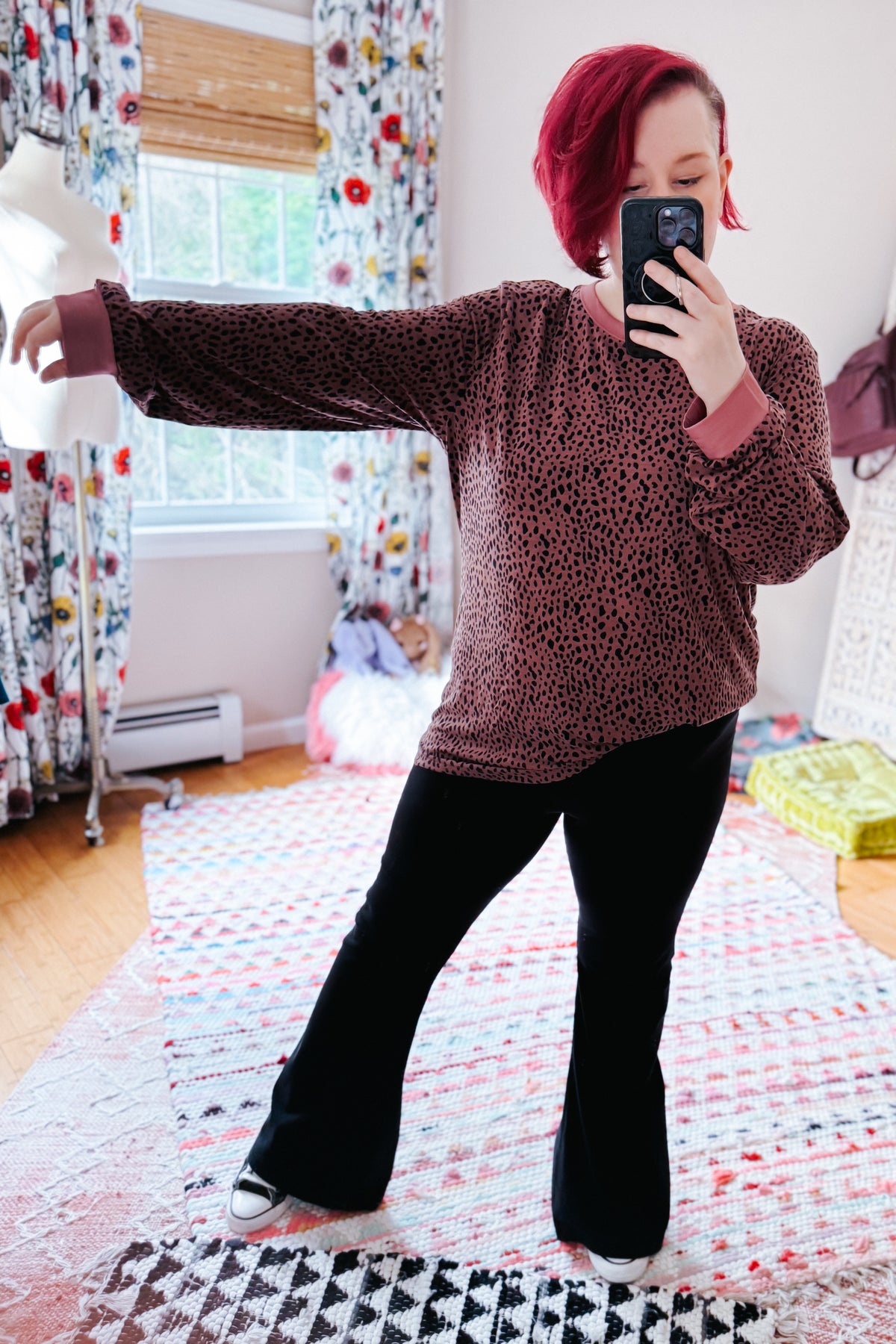 Red haired woman models a pink spotted long sleeve top with black flared pants and white sneakers during a bedroom mirror selfie