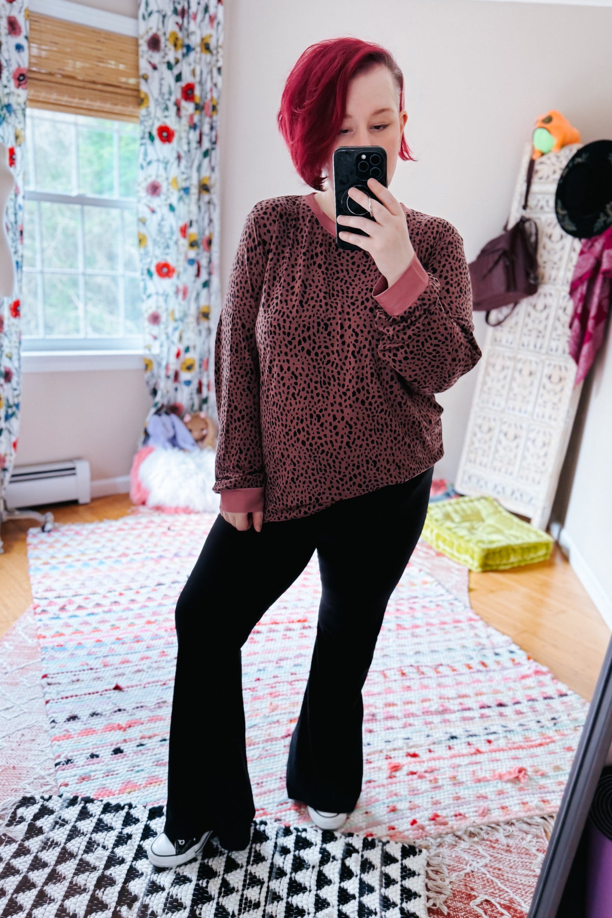 Short red haired woman models a pink spotted long sleeve top with black flared pants and white sneakers in a colorful bedroom