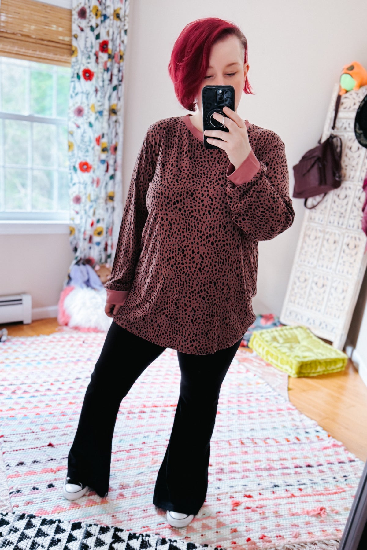 Short red haired woman models a pink spotted long sleeve top with black flared pants and white sneakers in a bright bedroom