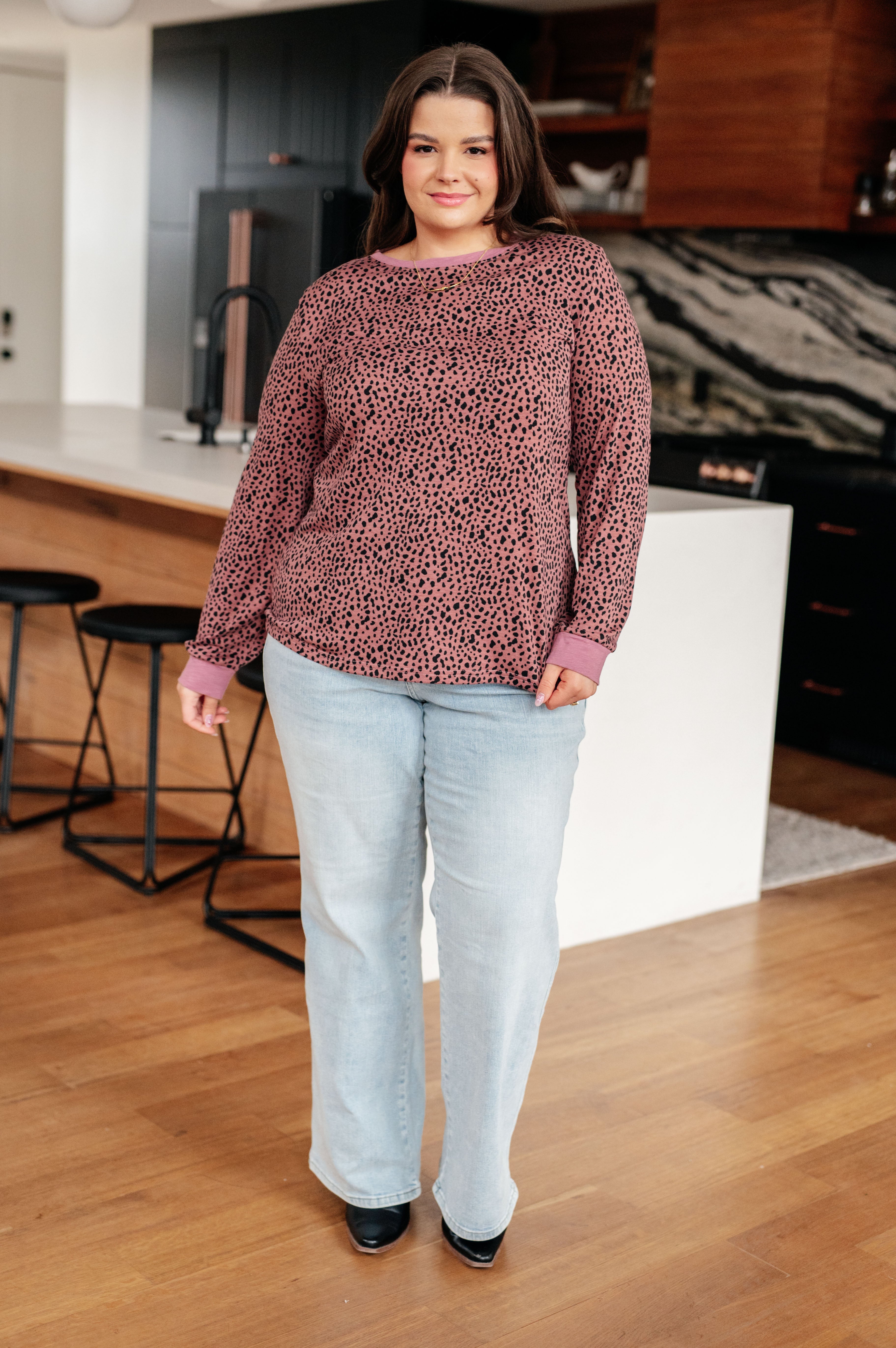 Plus-size woman wearing pink spotted long sleeve top paired with light-wash jeans in modern kitchen setting