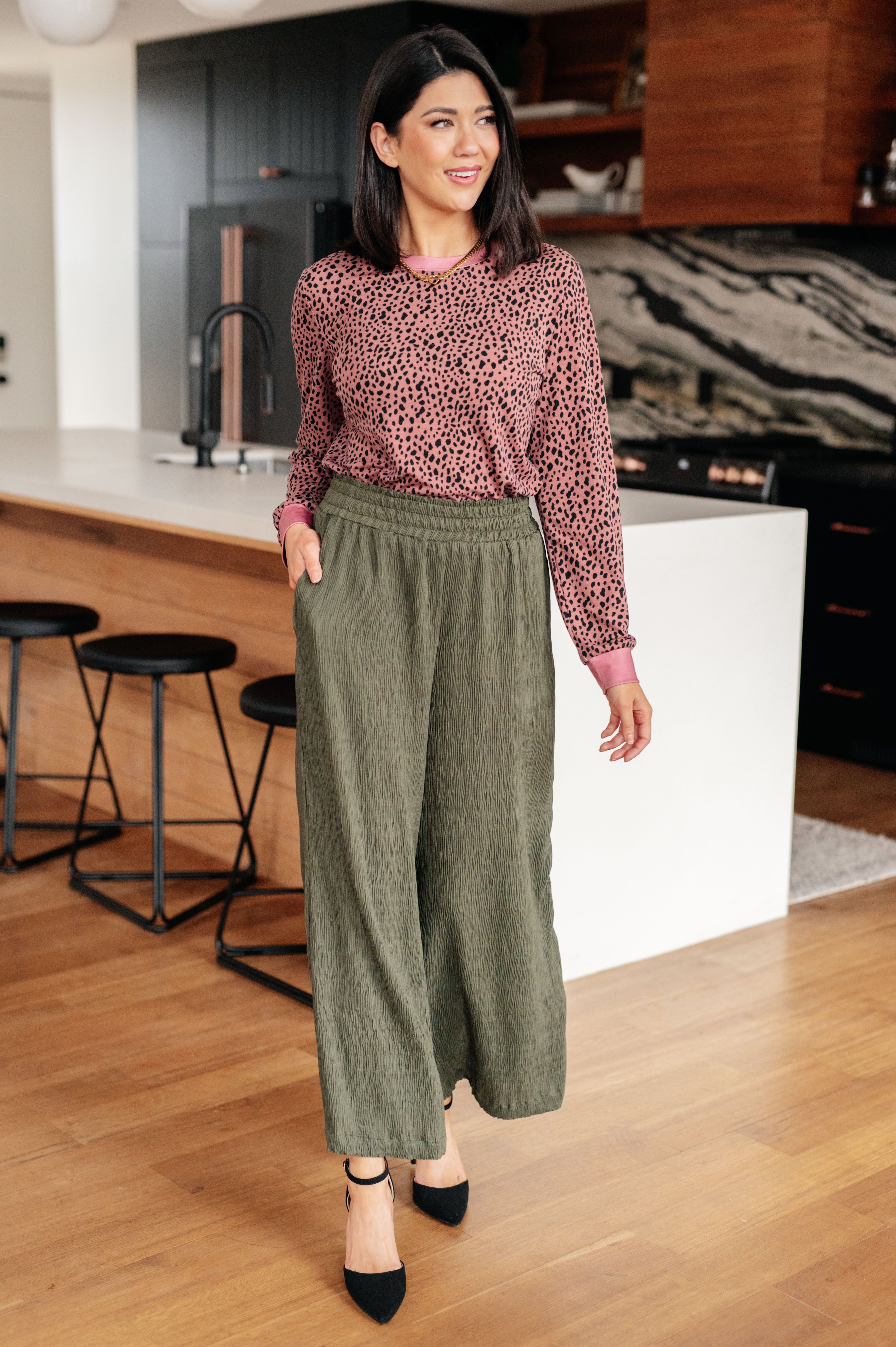 Woman wearing pink spotted long sleeve top with olive wide-leg pants walking in modern kitchen