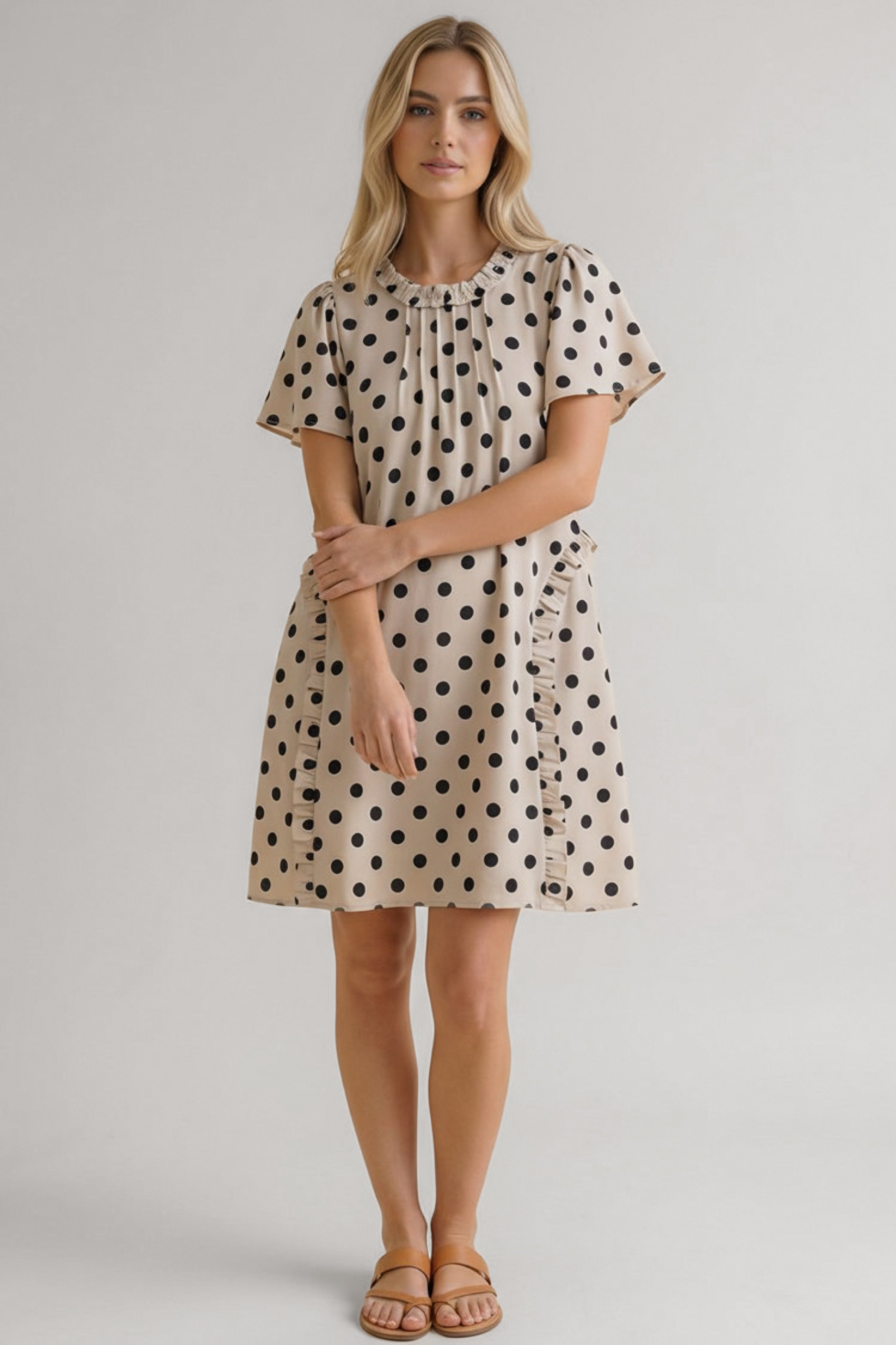 Woman wearing a beige dress with large black polka dots, short flutter sleeves, and ruffle trim along the front panel