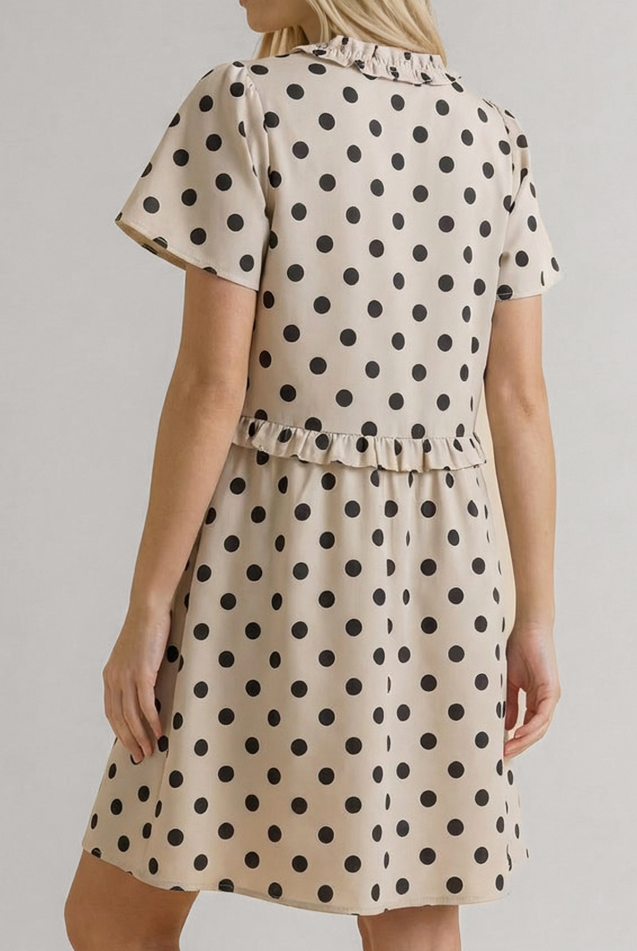 Woman in a beige dress with black polka dots, short sleeves, and ruffle details at the waist and collar viewed from the back