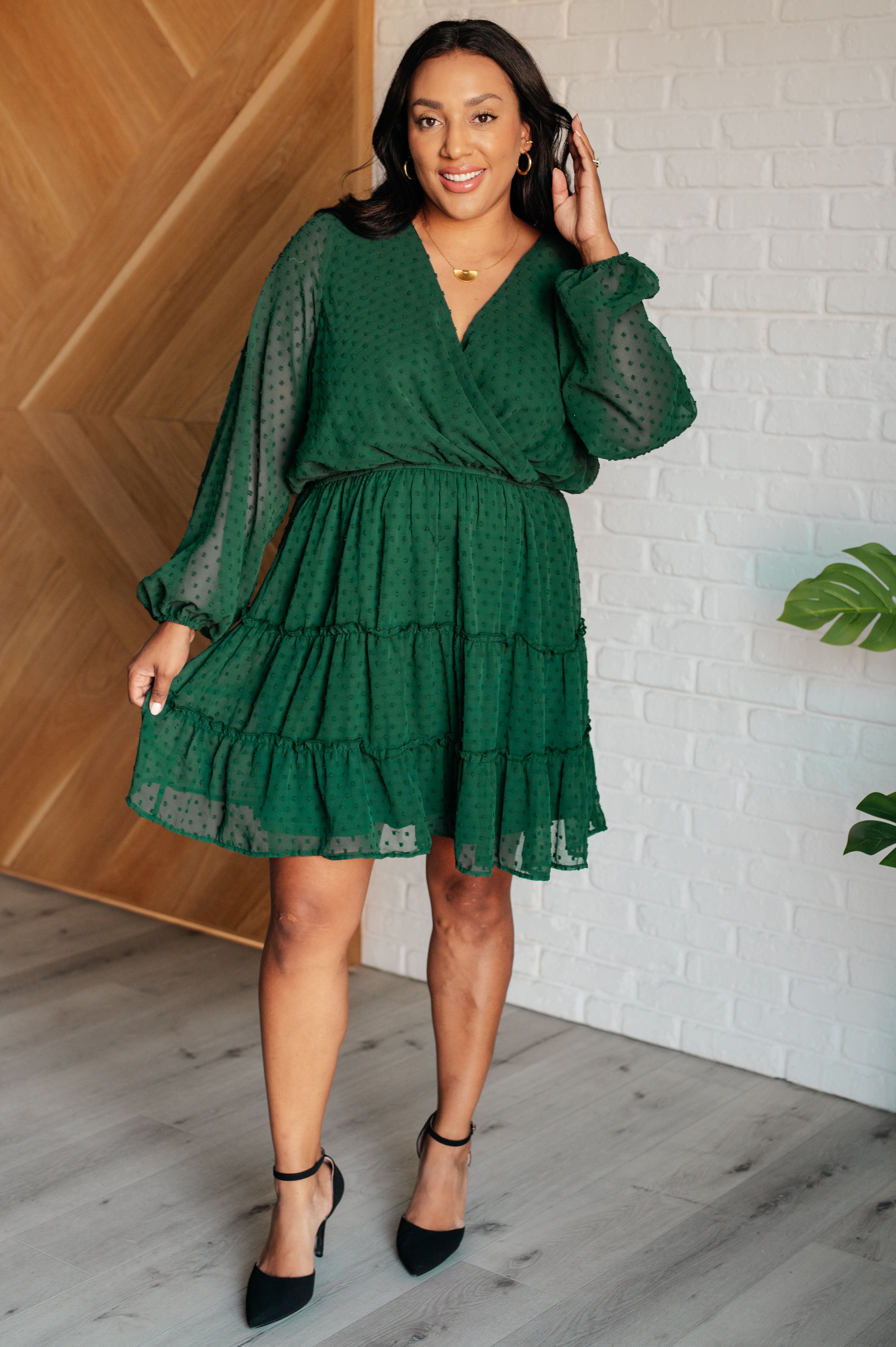 Woman wearing hunter green tiered dress with sheer long sleeves and textured dot pattern, styled with black heels indoors