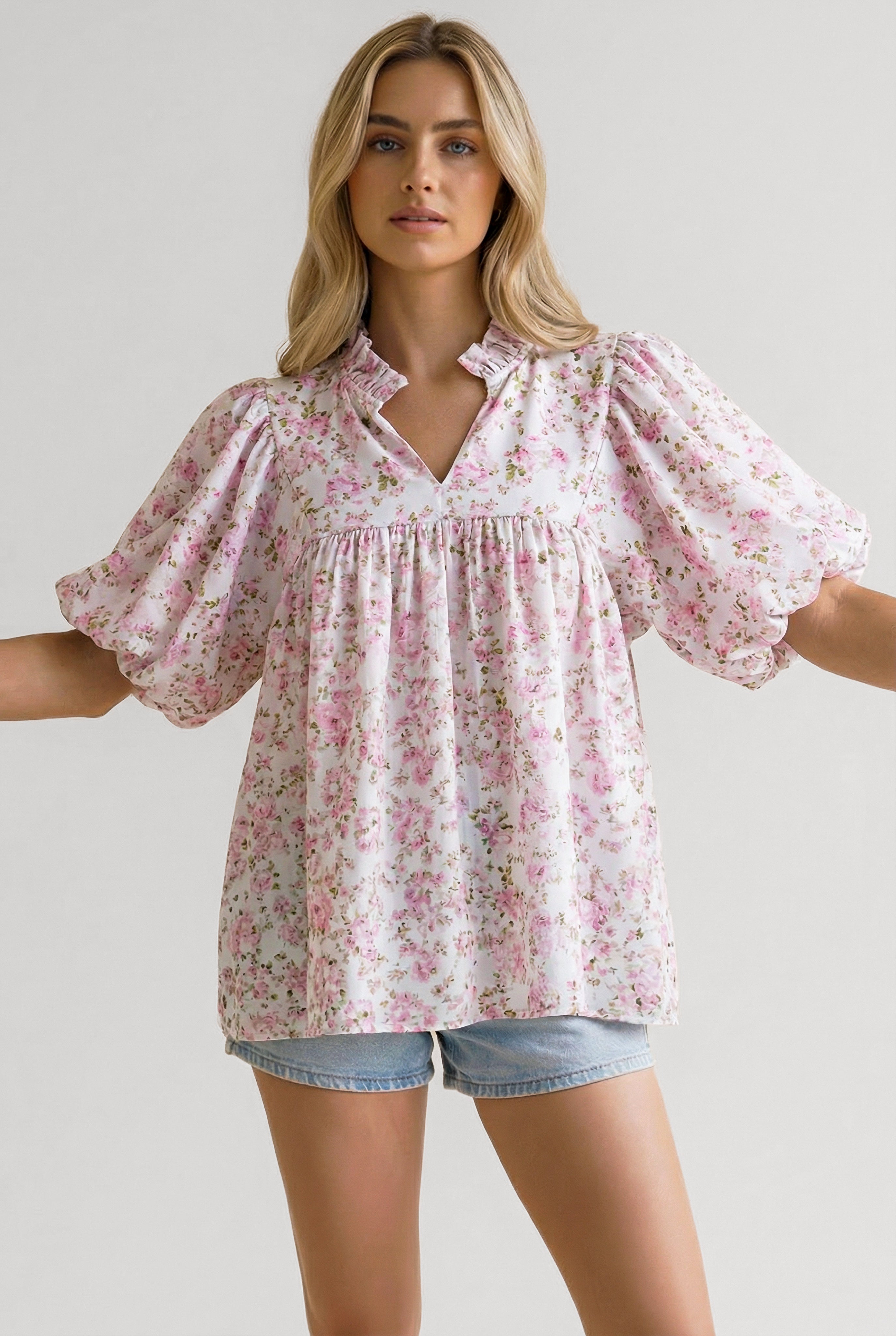 Woman wearing a loose pink floral blouse with puffed short sleeves and a ruffled neckline paired with light denim shorts