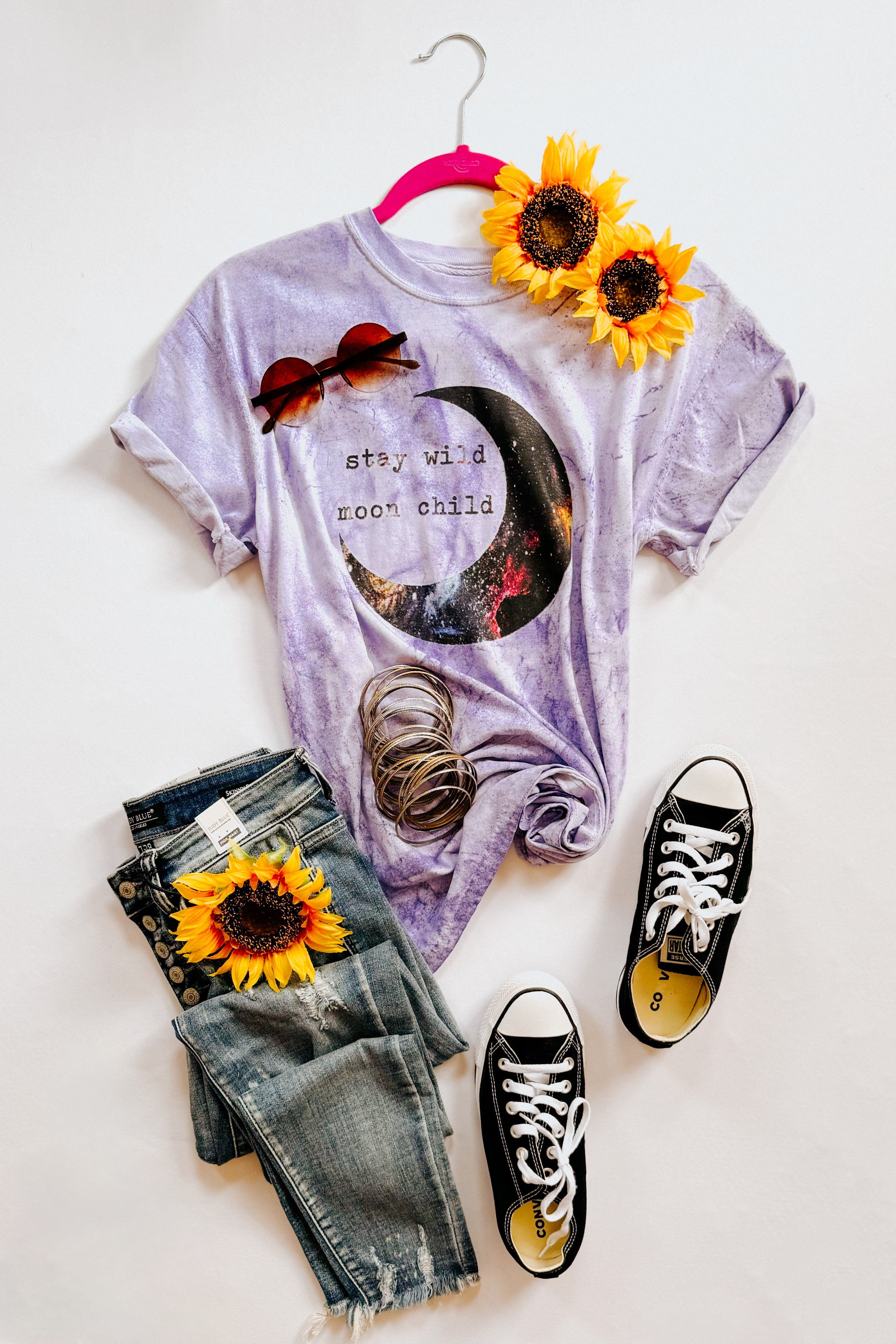 Purple graphic tee with moon print styled with black sneakers, distressed jeans, sunglasses, bracelets, and sunflowers on white background