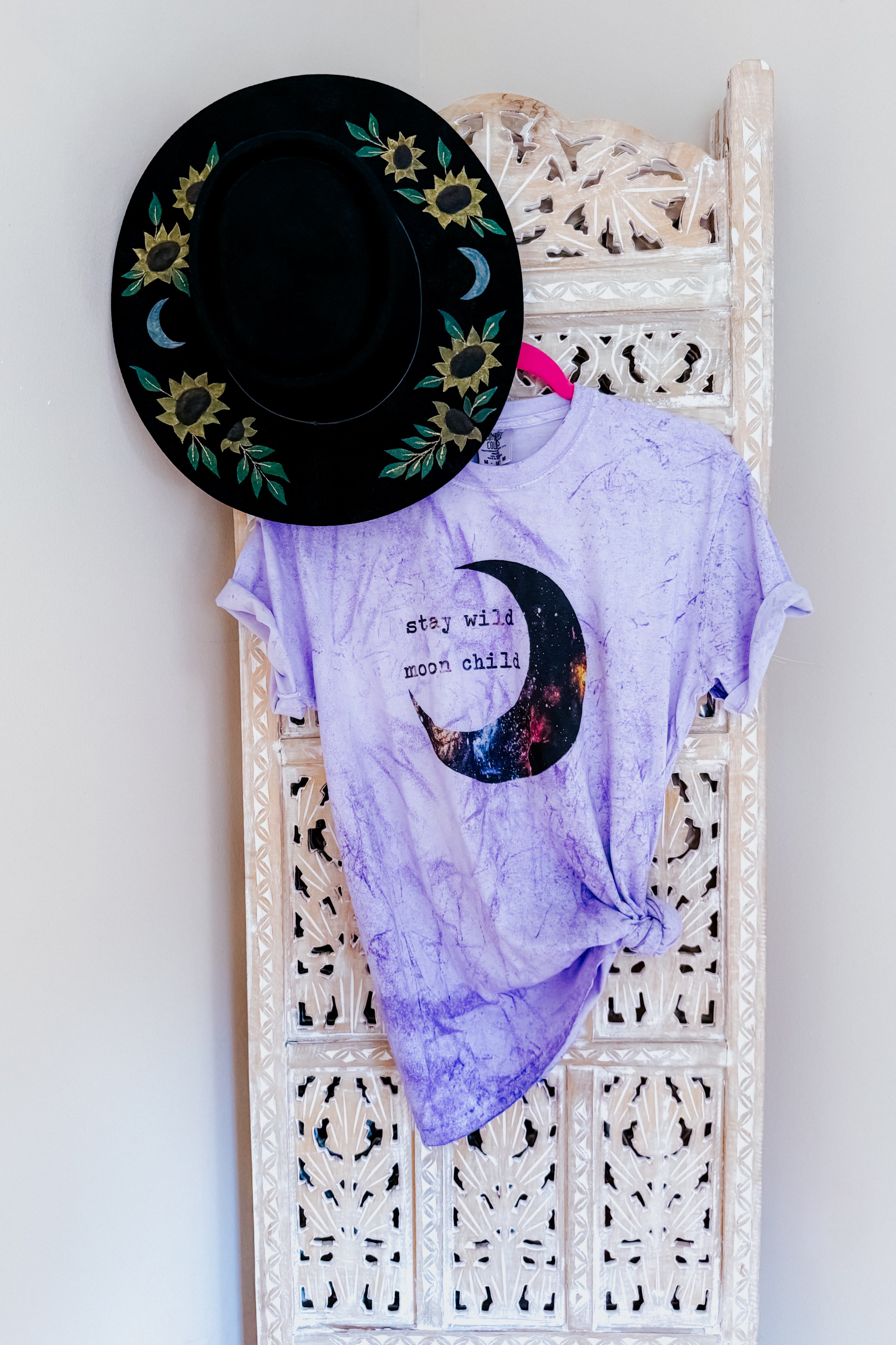 Purple tie-dye tee with crescent moon print and text "stay wild moon child" hanging next to black hat with floral designs