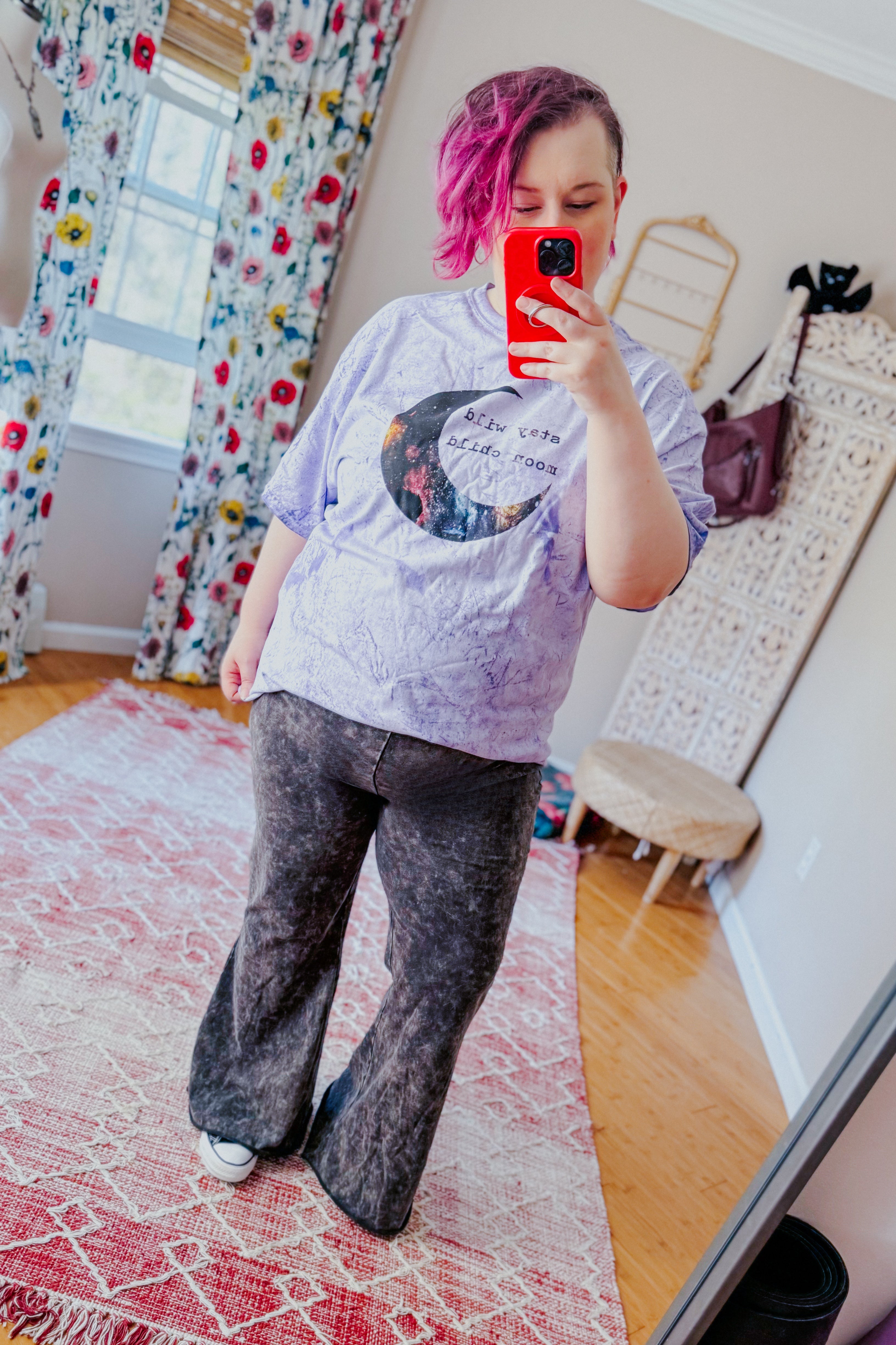 Woman with shaved side and pink hair wearing purple graphic tee and black acid wash wide-leg pants indoors