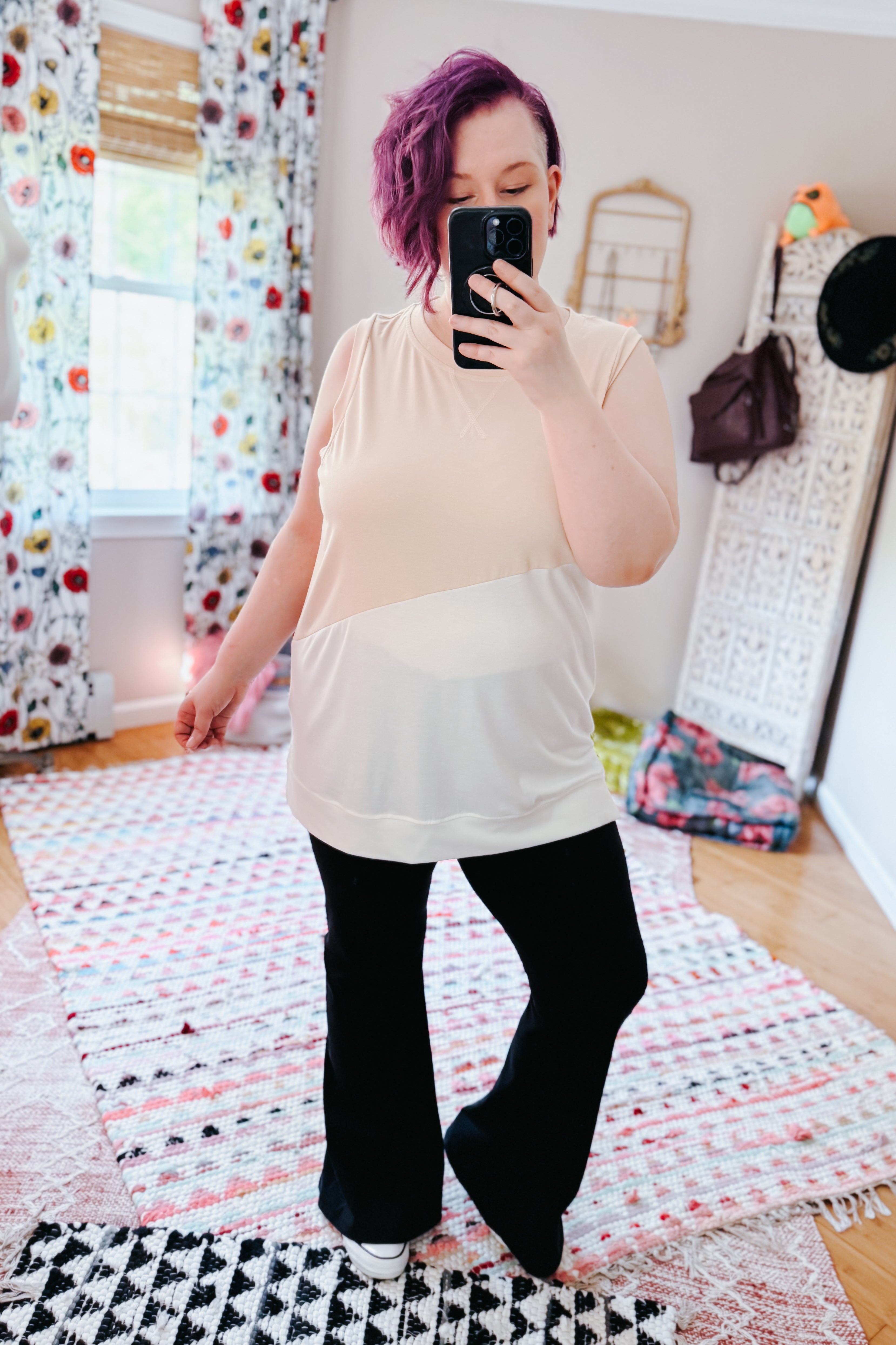 Woman with purple hair wearing a cream color block sleeveless top and black wide-leg pants in an eclectic, free-spirited room