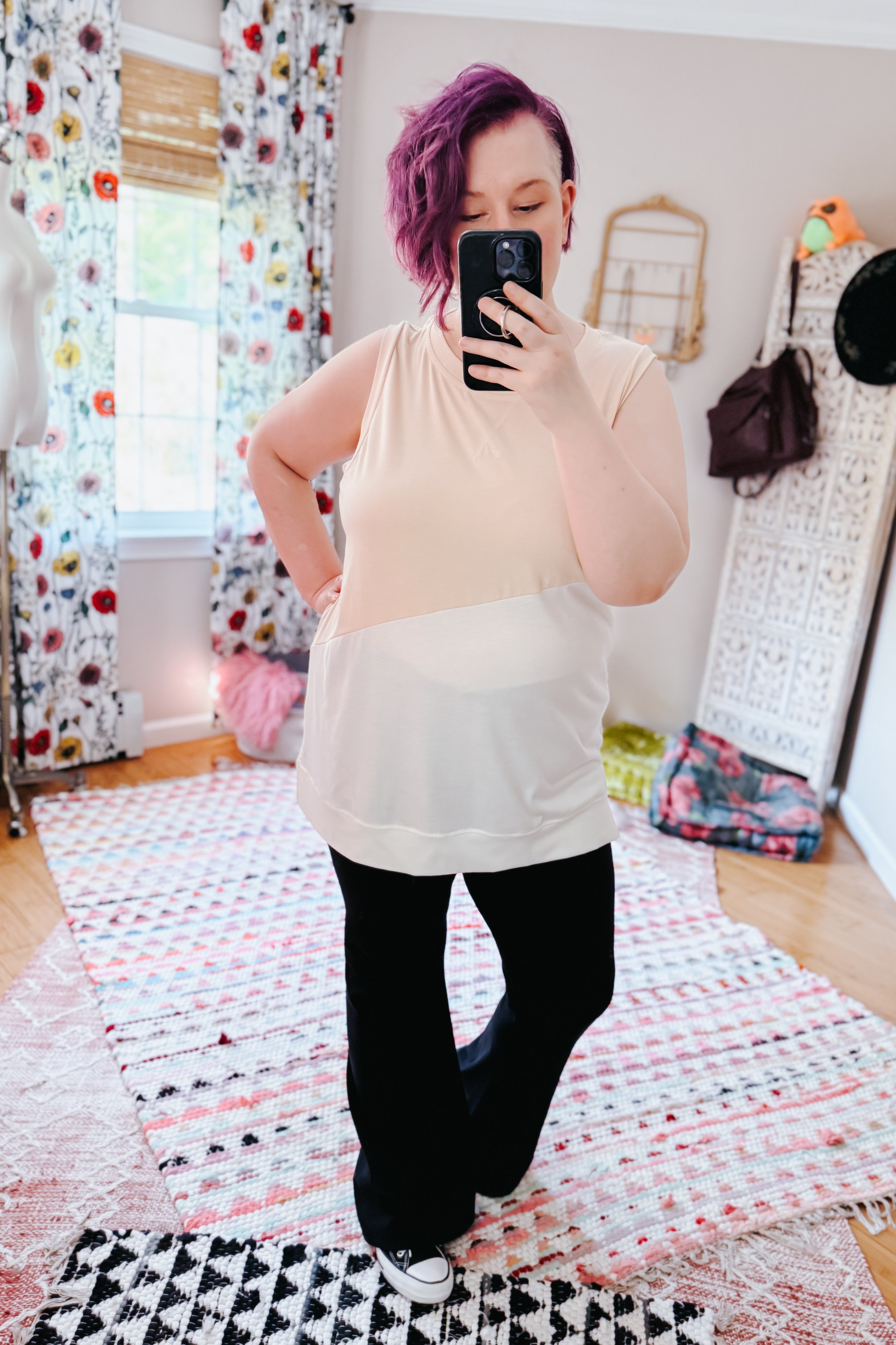 Woman with purple hair wearing a cream color block sleeveless top and black pants in a bright eclectic room