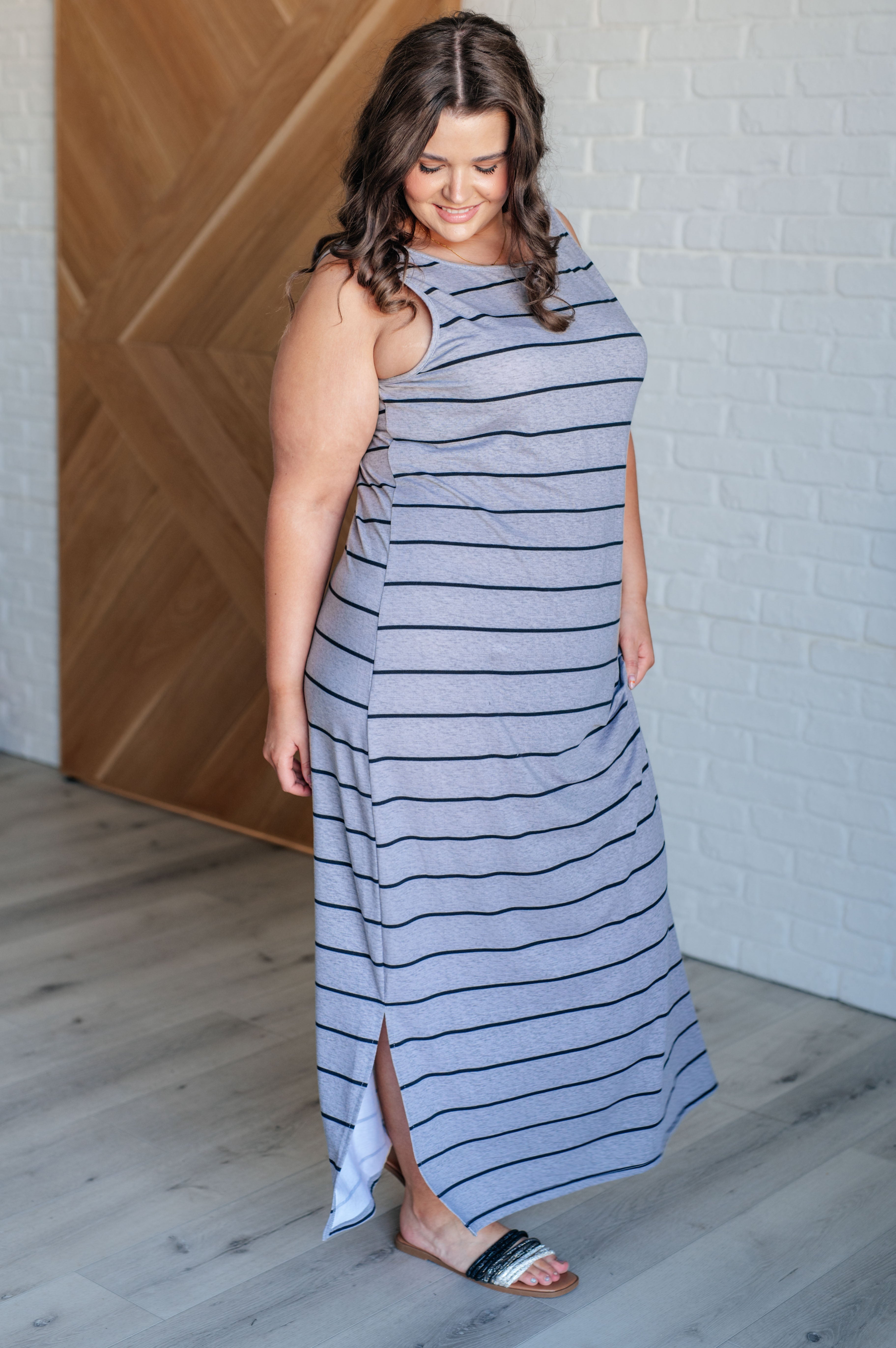 Woman wearing a gray sleeveless maxi dress with black stripes and side slit, paired with slide sandals indoors