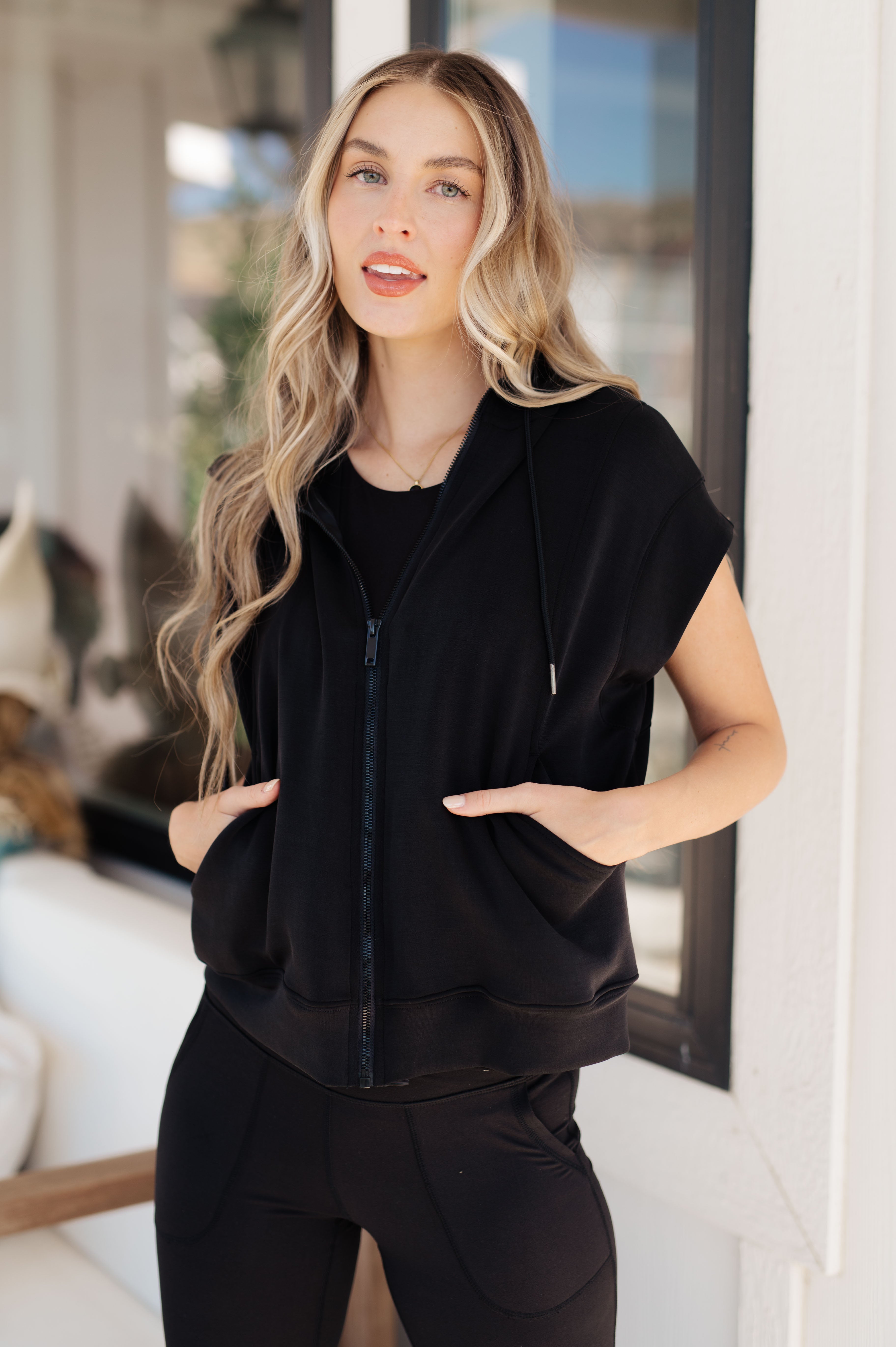 Woman with long wavy hair wearing a black sleeveless zip hoodie and matching pants standing by window