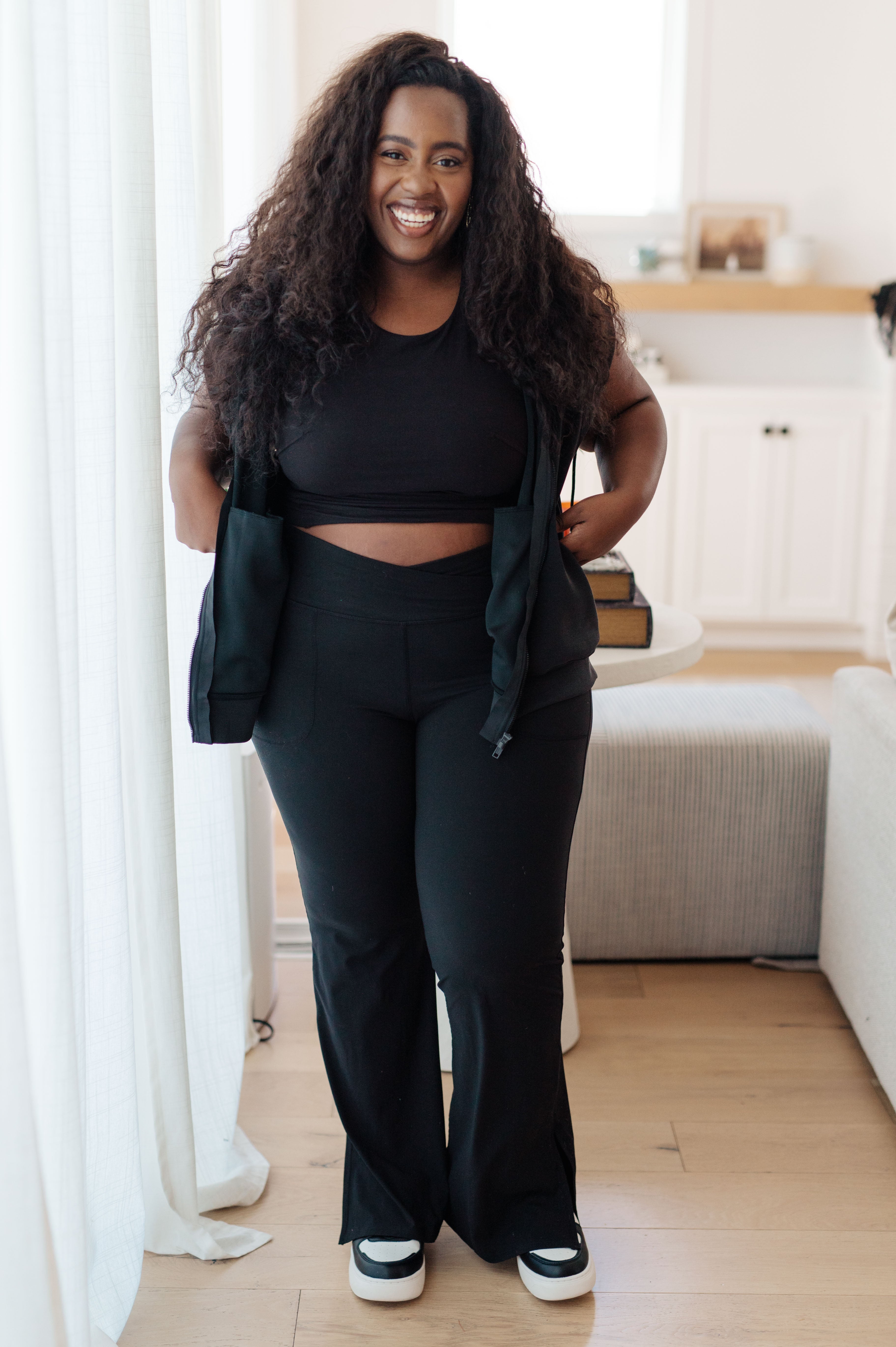 Smiling plus-size woman wearing black sleeveless hoodie, black crop top, and black pants standing indoors by white curtain
